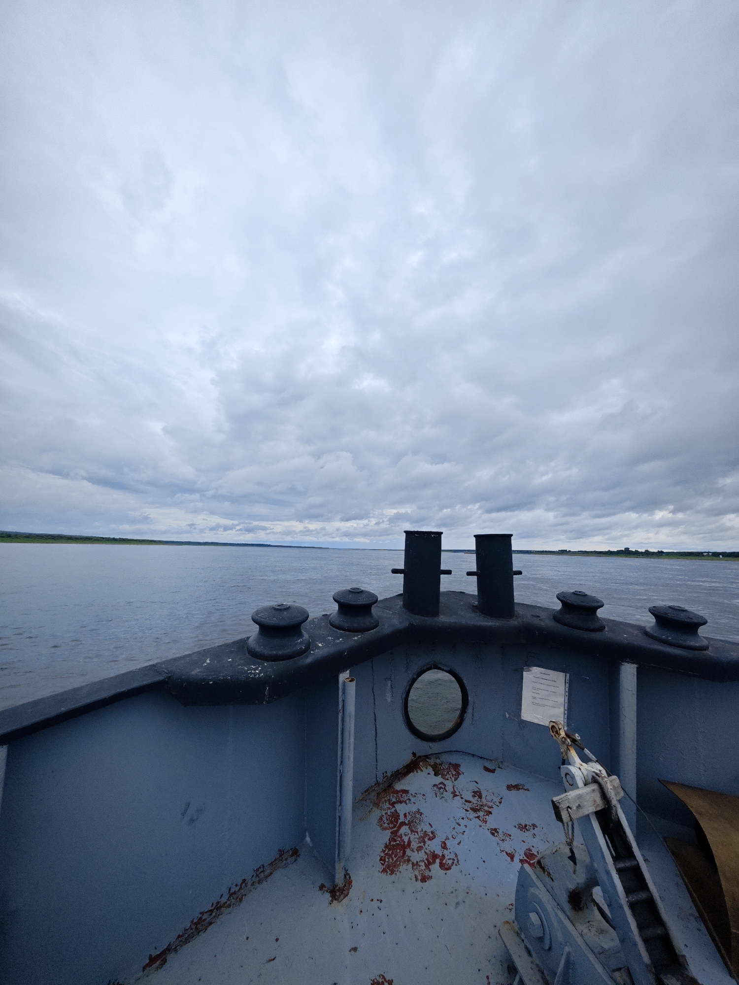 The image is a low-angle, wide-shot perspective taken from the bow of a gray boat on a large body of water, likely a river given the flat landscape on the horizon. The sky is overcast with a thick layer of gray clouds. The water is calm and reflects the muted light.
In the foreground, the deck of the boat is visible. It is painted gray and shows signs of wear and tear, with patches of rust-colored material and peeling paint. Two dark, rounded bollards are prominent on the deck, one in the middle ground left and another to its right. Further back on the deck, two taller, black cylindrical structures, likely cleats or mooring posts, stand side-by-side. Between these, a circular porthole is visible on the boat's structure, looking out onto the water. A white document is affixed to the wall near the porthole. To the right of the porthole and cleats, a mechanism with metal and wooden components, possibly part of the boat's anchor system, is visible.
The horizon is defined by a thin strip of green land, suggesting a distant shoreline or island. The overall impression is one of overcast weather on a calm waterway, from the perspective of someone on the vessel. The location is Zotino, Russia.