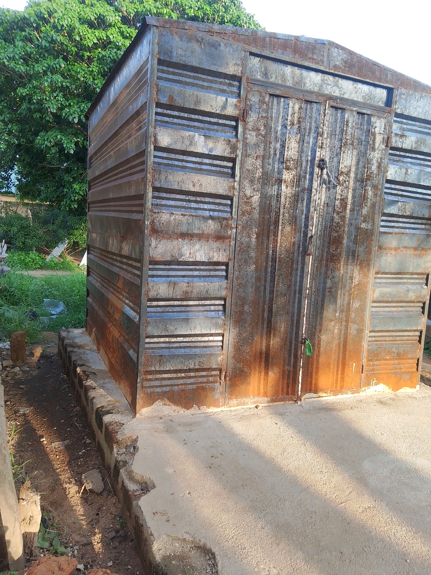 This image captures an outdoor scene in Jalingo, Nigeria, featuring a rusted corrugated metal shed with double doors. The shed is situated on a concrete foundation with a brick border. Lush green foliage and trees are visible in the background, suggesting a semi-rural or suburban setting. The lighting indicates it is daytime, with shadows cast across the concrete. The shed appears weathered, with visible rust and signs of age.