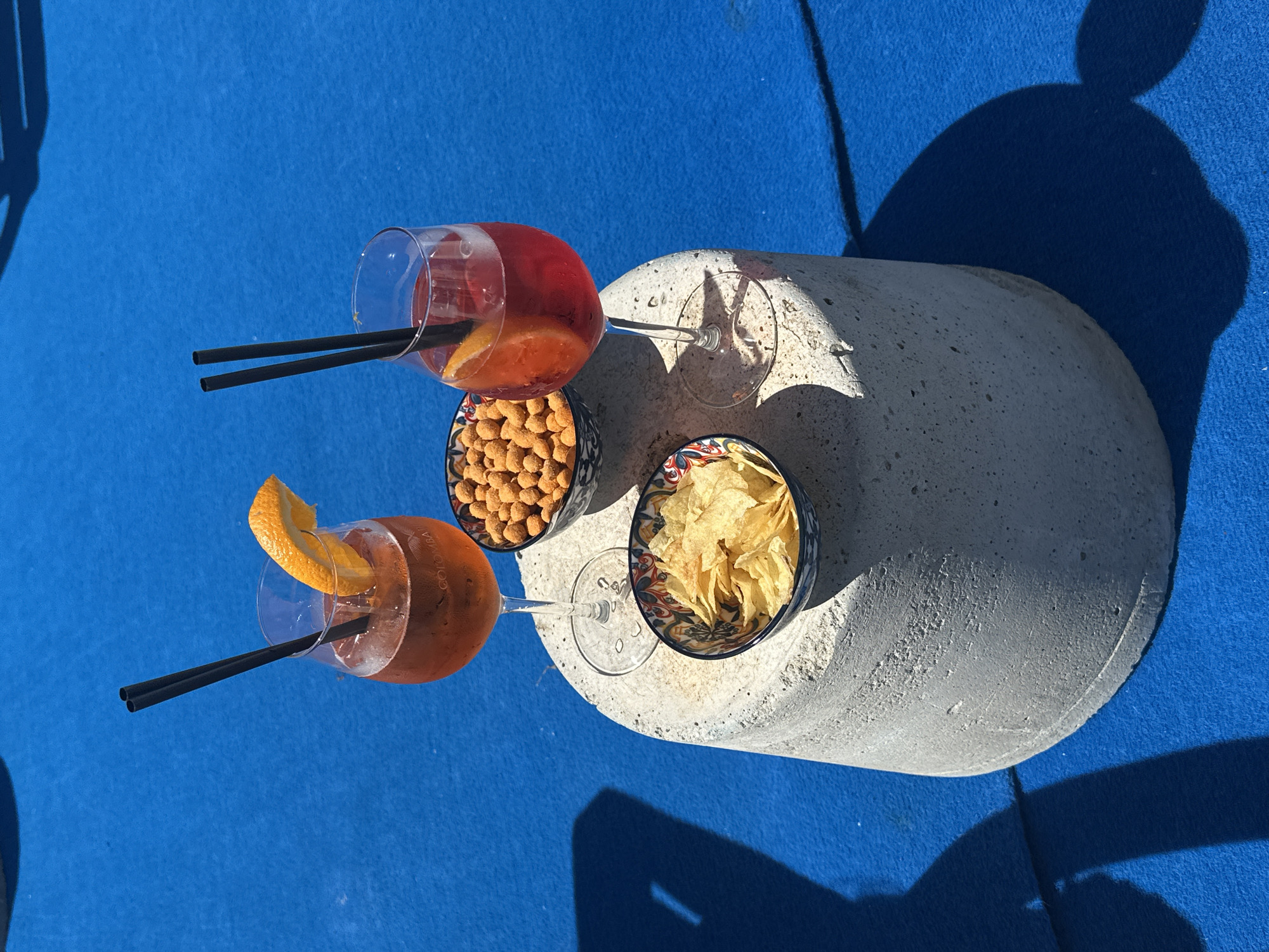 The image presents a vibrant, top-down view of an outdoor refreshment setup, likely at a resort or by a pool. Two cocktails are prominently featured, each served in a stemmed glass with an orange slice garnish and two black straws. The drinks are a bright orange-red hue, suggesting Aperol Spritz or similar summer cocktails. One glass faintly displays the text "COLOMBA".
Accompanying the beverages are two small, patterned bowls of snacks: one filled with roasted peanuts or similar nuts, and the other with potato chips. These items are arranged on a curved, light gray, textured concrete surface, which contrasts with the deep blue, textured fabric background, likely a sunbed or towel.
The scene is bathed in bright, direct sunlight, casting sharp, distinct shadows from the glasses, the concrete surface, and unseen objects or individuals to the left and top of the frame. This indicates it is daytime, likely midday or early afternoon, under clear skies. The overall impression is one of relaxation and leisure, typical of a vacation setting in a sunny location such as Lipari, Italy. No people are visible in the frame, but their presence is implied by the drinks and snacks.