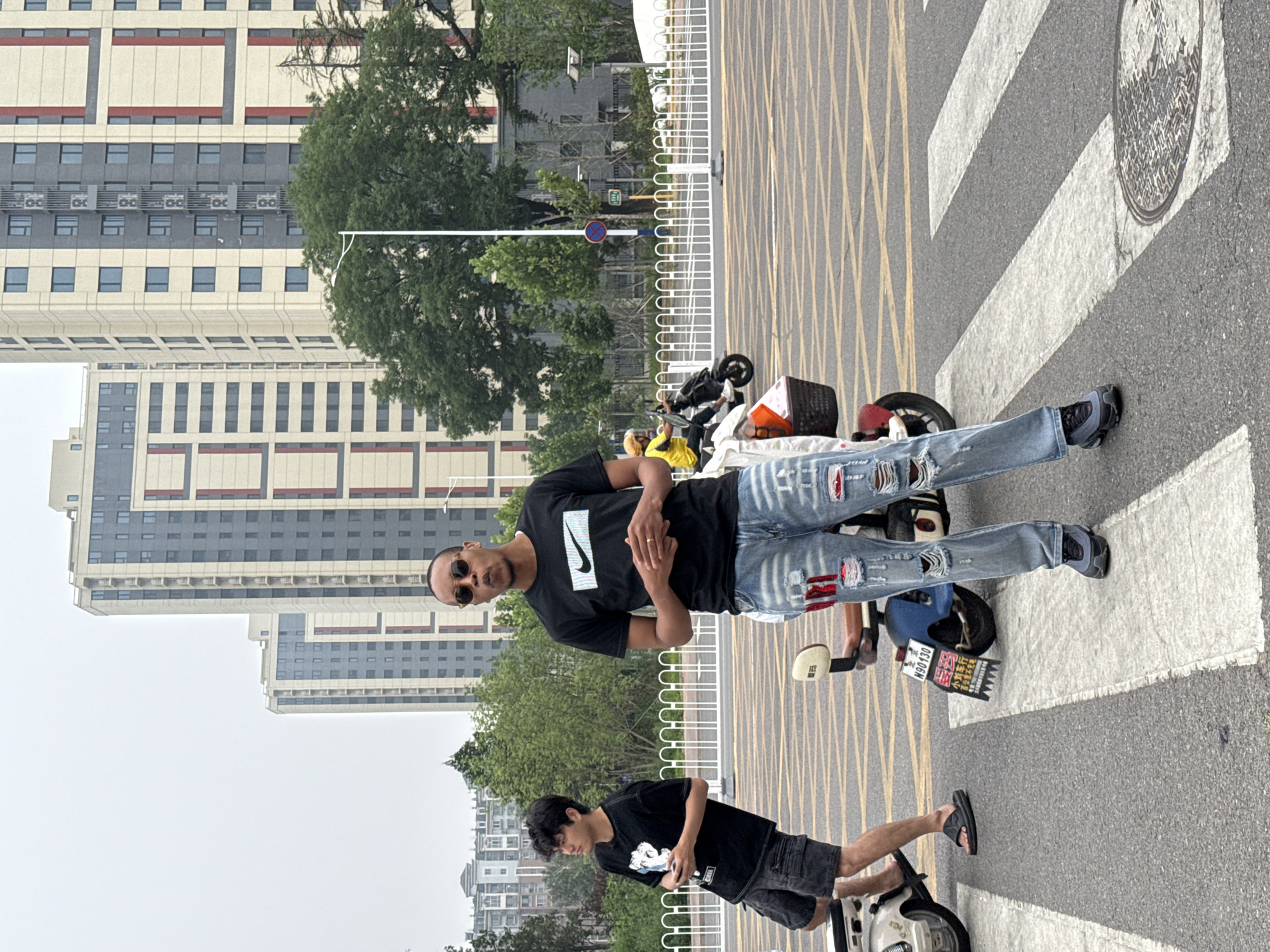 Two young men are standing on a crosswalk in the Daxing District of China. The man on the right is wearing a black Nike t-shirt and ripped blue jeans, and sunglasses. He is looking forward with his mouth open. The man on the left is wearing a black t-shirt and denim shorts, and is looking to the left. Two electric scooters are parked behind them. In the background, there are tall, modern buildings and trees. The sky is overcast.