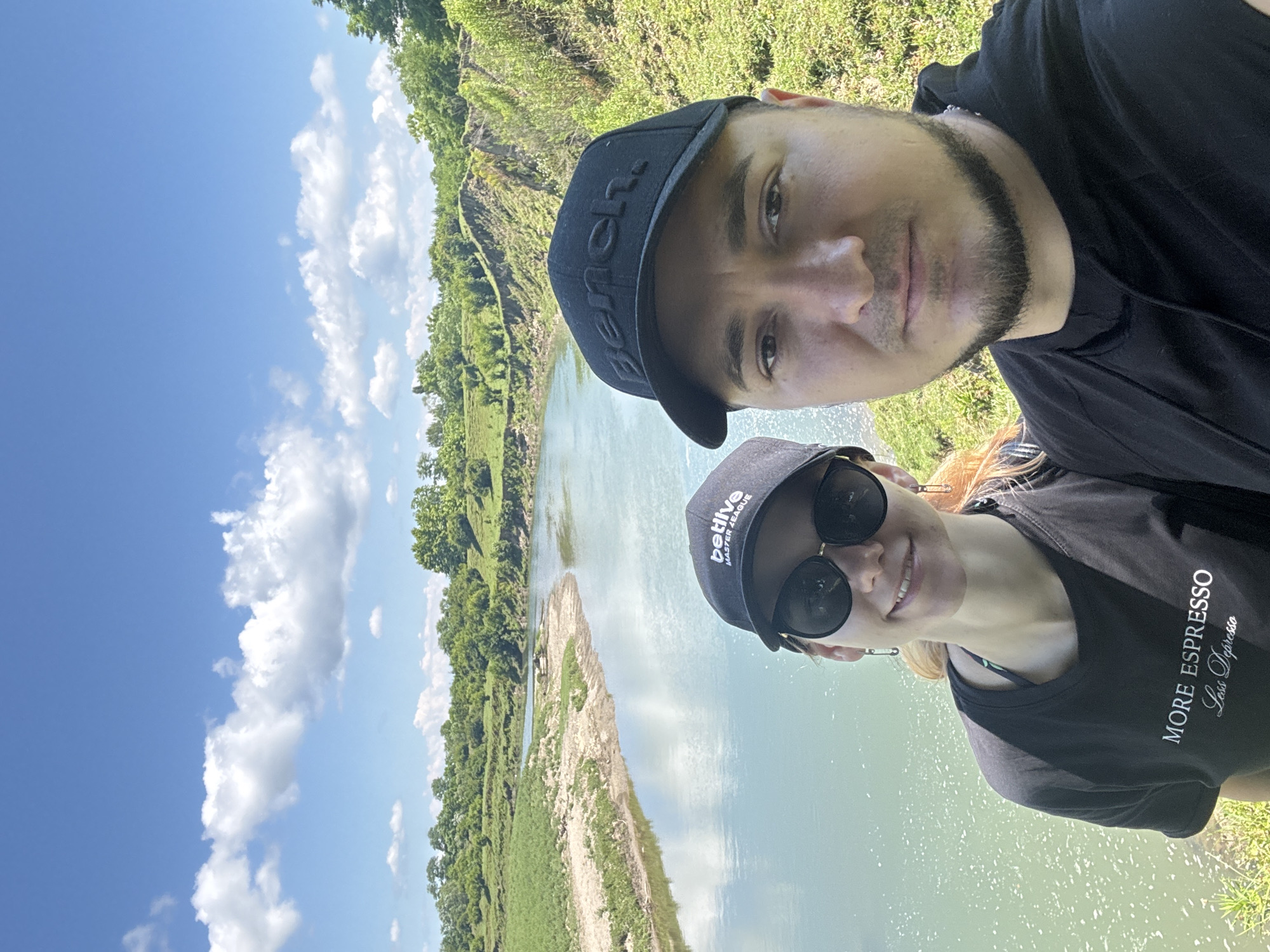 An outdoor selfie is taken by a couple standing near a river under a blue sky with scattered white clouds. The man on the right wears a black baseball cap and a black hoodie. The woman on the left wears a black baseball cap, sunglasses, and a black t-shirt with the text "MORE ESPRESSO Less Depresso" printed on it. She is also wearing distinctive safety pin earrings. The river, which appears to be a light blue-green color, flows from the left to the right of the frame. The banks of the river are lined with green vegetation, and in the background, there are rolling hills covered in trees and shrubs. The landscape suggests a natural, possibly rural, setting. The location is identified as Darcheli, Georgia.
