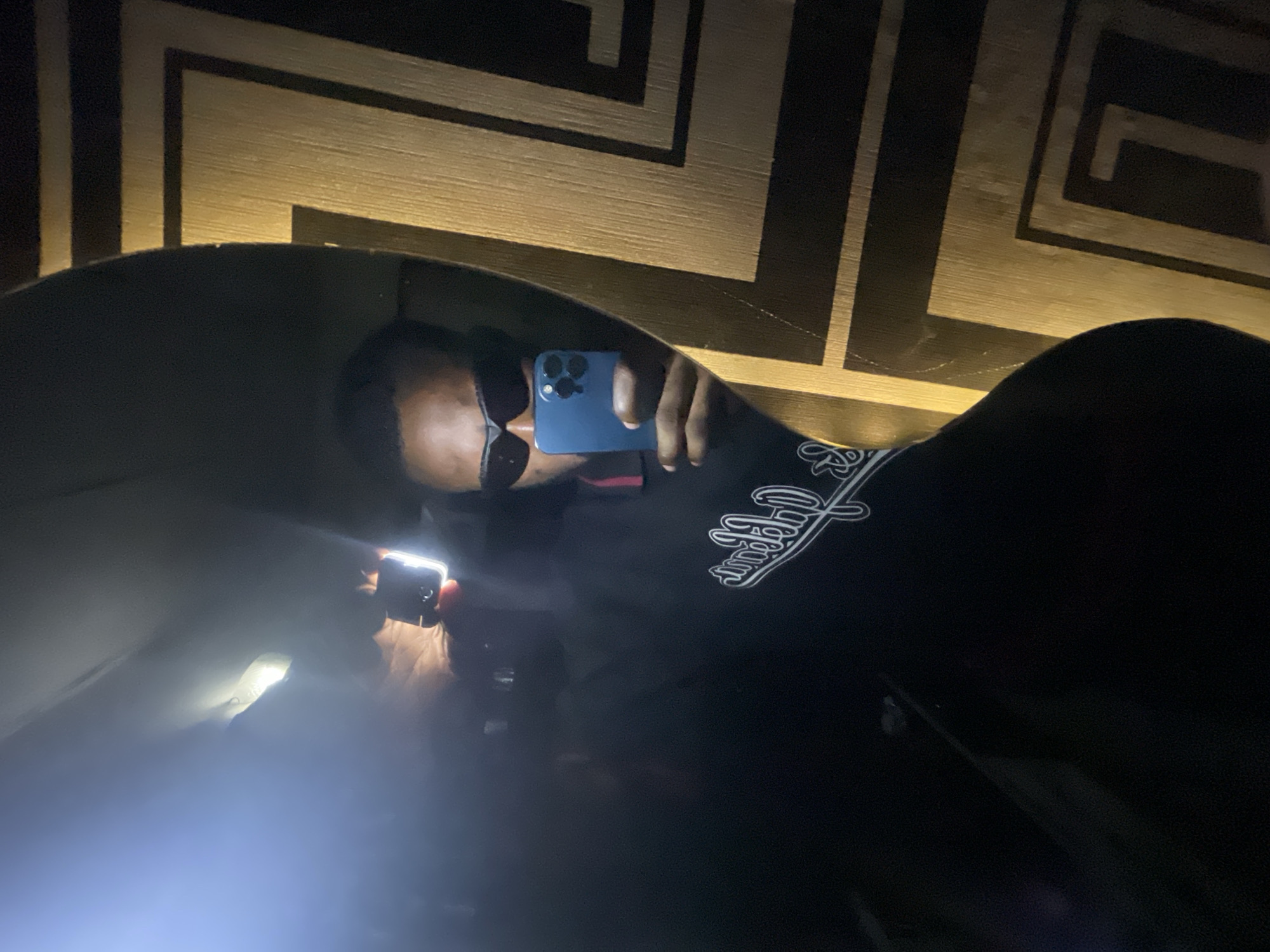 A person wearing sunglasses is taking a mirror selfie in Lokoja, Nigeria. The person is holding a blue iPhone in their right hand, and a bright light from the camera flash is illuminating the scene. The background appears to be a wall with a geometric pattern. The person is wearing a black sweatshirt with white script writing on the left chest. The overall lighting of the image is dim, with the flash creating a strong contrast.