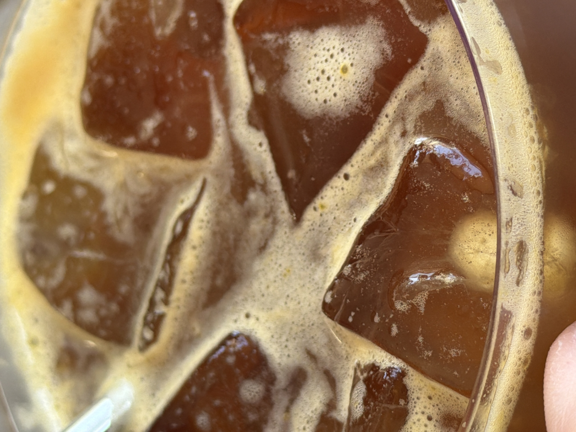 An overhead shot of a glass filled with ice and a dark liquid, likely iced coffee or tea, with a straw visible in the lower-left corner. The glass rim is visible on the right. Small bubbles are present on the surface of the liquid and around the ice. Part of a human finger is visible in the lower right corner. The image provides no direct information about the location in Bishkek City, Kyrgyzstan.