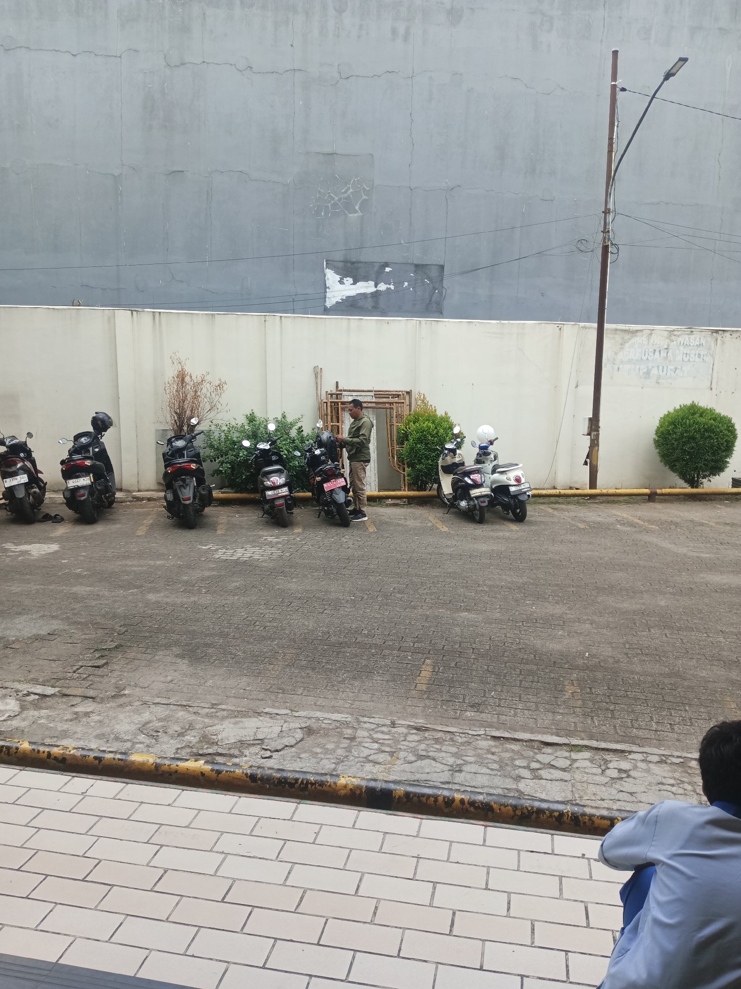 The image depicts an outdoor parking area in South Tangerang, Indonesia. Several motorcycles are parked in a row against a light-colored wall. A man in a green shirt and khaki pants is standing next to one of the motorcycles, appearing to interact with it. The ground is paved with textured blocks, and a yellow and black striped barrier runs along the edge of the parking lot. In the background, a tall, grey wall with visible cracks dominates the scene, with a utility pole and electrical wires. To the right, a large green, rounded bush is situated. In the foreground, a person in a blue shirt and pants is seated facing away from the camera. The overall impression is one of a typical urban environment.