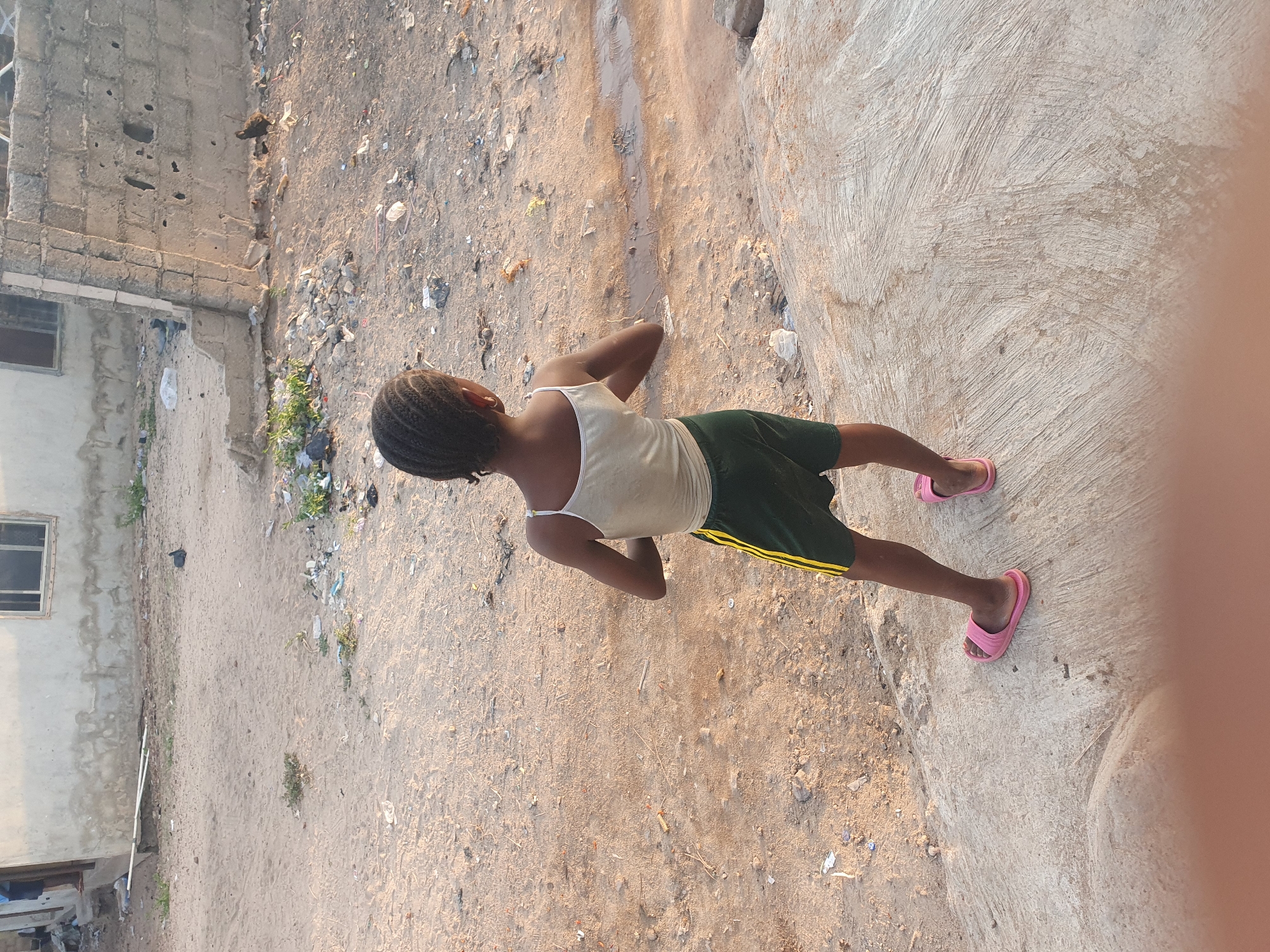 An overhead photograph captures a young person, likely a girl, standing outdoors in Lokoja, Nigeria. The individual, with dark skin and braided hair, is viewed from behind. She wears a light-colored (possibly white or beige) tank top and dark green shorts featuring two yellow stripes along the outer seams. Pink slide sandals are visible on her feet. Her arms are bent at the elbows, with her hands resting behind her lower back as she gazes towards her right (the viewer's left).
The setting appears to be a semi-urban or residential area. The ground surface is varied, transitioning from a sandy, dirt-covered section on the left, which is strewn with small debris and sparse weeds, to a rough concrete path or slab on the right. A simple building constructed of concrete blocks, featuring a single window, is visible on the far left edge of the frame. The lighting indicates either late afternoon or early morning, with warm, soft sunlight casting gentle shadows, suggesting clear weather. No specific activity is depicted beyond the individual standing and observing the surroundings. There is no visible text in the image.