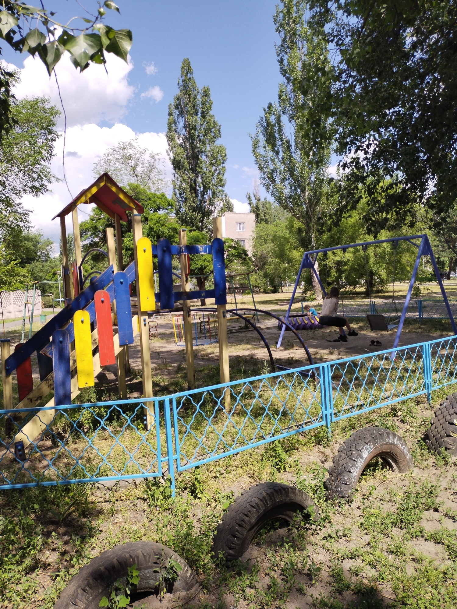 This is a medium-angle, eye-level outdoor photograph of a playground in Horishni Plavni, Ukraine. The playground features a multi-colored wooden climbing structure with blue, yellow, and red elements. To the right of the climbing structure, there is a blue swing set with two swings. A young person is seated on one of the swings. The playground is enclosed by a blue metal fence with a diamond pattern. In the foreground, several old tires are partially buried in the ground, overgrown with vegetation. Behind the playground, there are trees and a multi-story building. The sky is blue with scattered white clouds. The overall condition of the playground appears weathered and somewhat neglected.