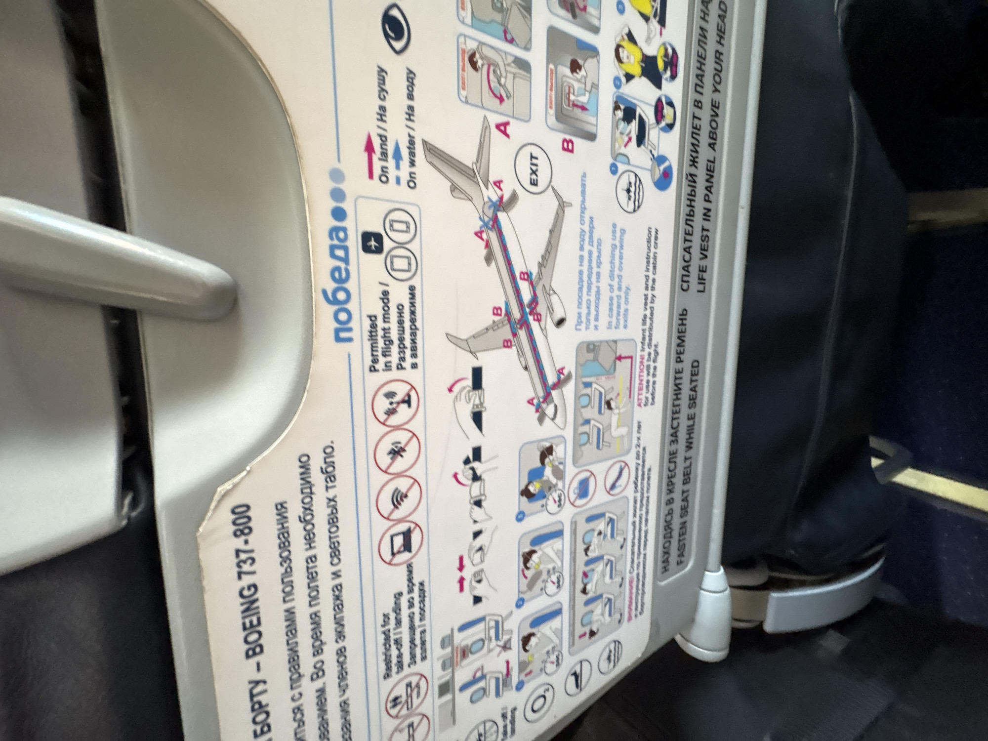 This is a photograph taken inside an airplane cabin. The focus is on the safety instruction card affixed to the seat in front. The card is labeled with "BOEING 737-800" and the airline name "победа...". It displays pictograms and text instructions in Russian and English concerning safety procedures for passengers, including the use of electronic devices, emergency exits, and life vests. The seat and part of the adjacent seat can be seen around the card.