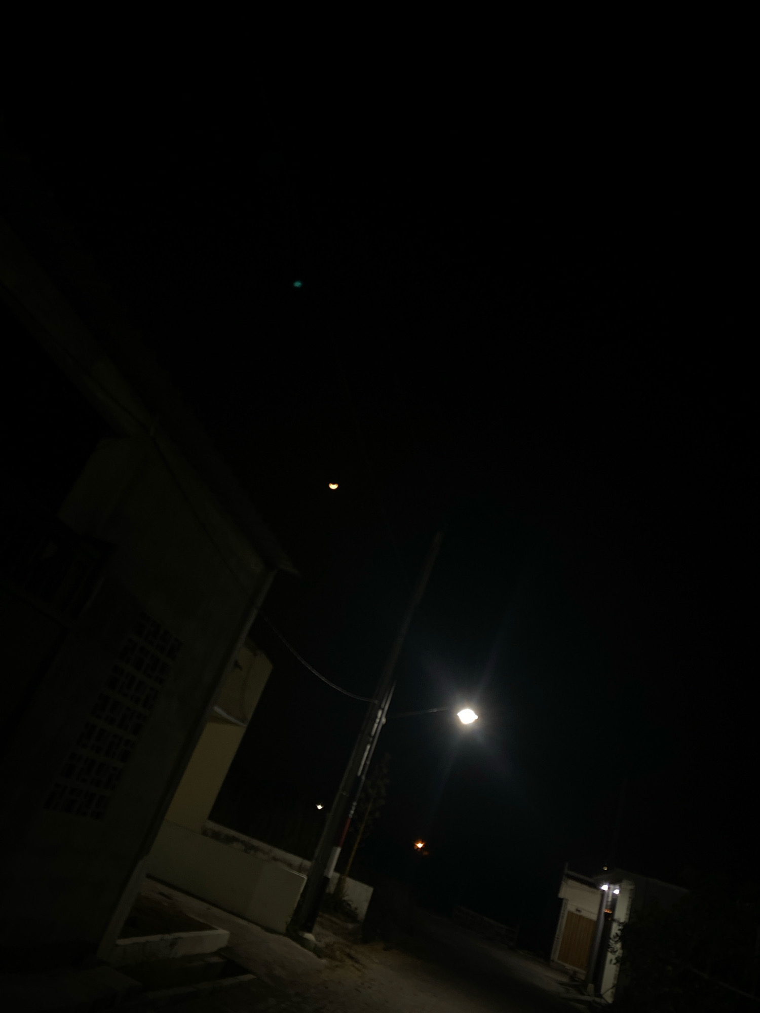 This is a nighttime photograph, possibly taken in Kedung Sukodani, Indonesia, depicting a street scene. The sky is dark with a crescent moon visible. A building with a textured wall and windows is partially visible on the left side of the frame. A street lamp illuminates a section of the unpaved street in the center-right. In the background, another building with a light fixture is visible. The image is dark overall, with limited detail due to the low light conditions.