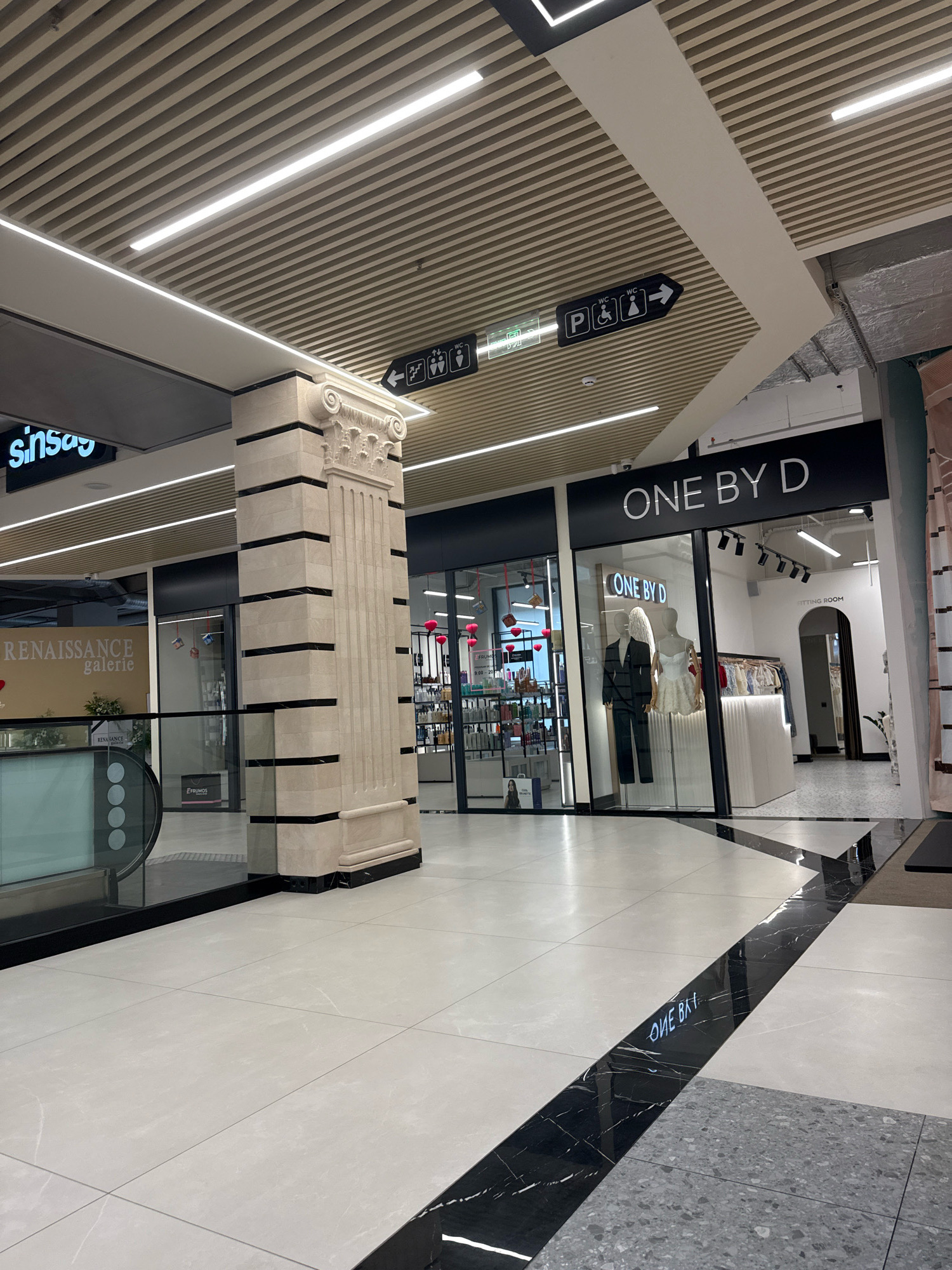 The image captures an interior view of a modern shopping mall. In the foreground, a polished floor with a striking black and white marble inlay leads the eye towards the background. To the left, a "Sinsay" store is partially visible, with an escalator also present. In the center, a large decorative column, made of stone with black horizontal banding and a classical capital, stands prominently. To the right of the column, a shop with the prominent signage "ONE BY D" is displayed, featuring mannequins in formal wear and a rack of clothing. Further back, a sign indicates "FITTING ROOM". Overhead, a grid-like ceiling with integrated LED lighting is visible. Signage indicating restrooms and accessibility is also present on the ceiling. The overall impression is of a clean, well-lit, and contemporary retail environment. Given the presence of "Sinsay" and "ONE BY D," it is likely this scene is located in the Shopping MallDova in Chișinău, Moldova.