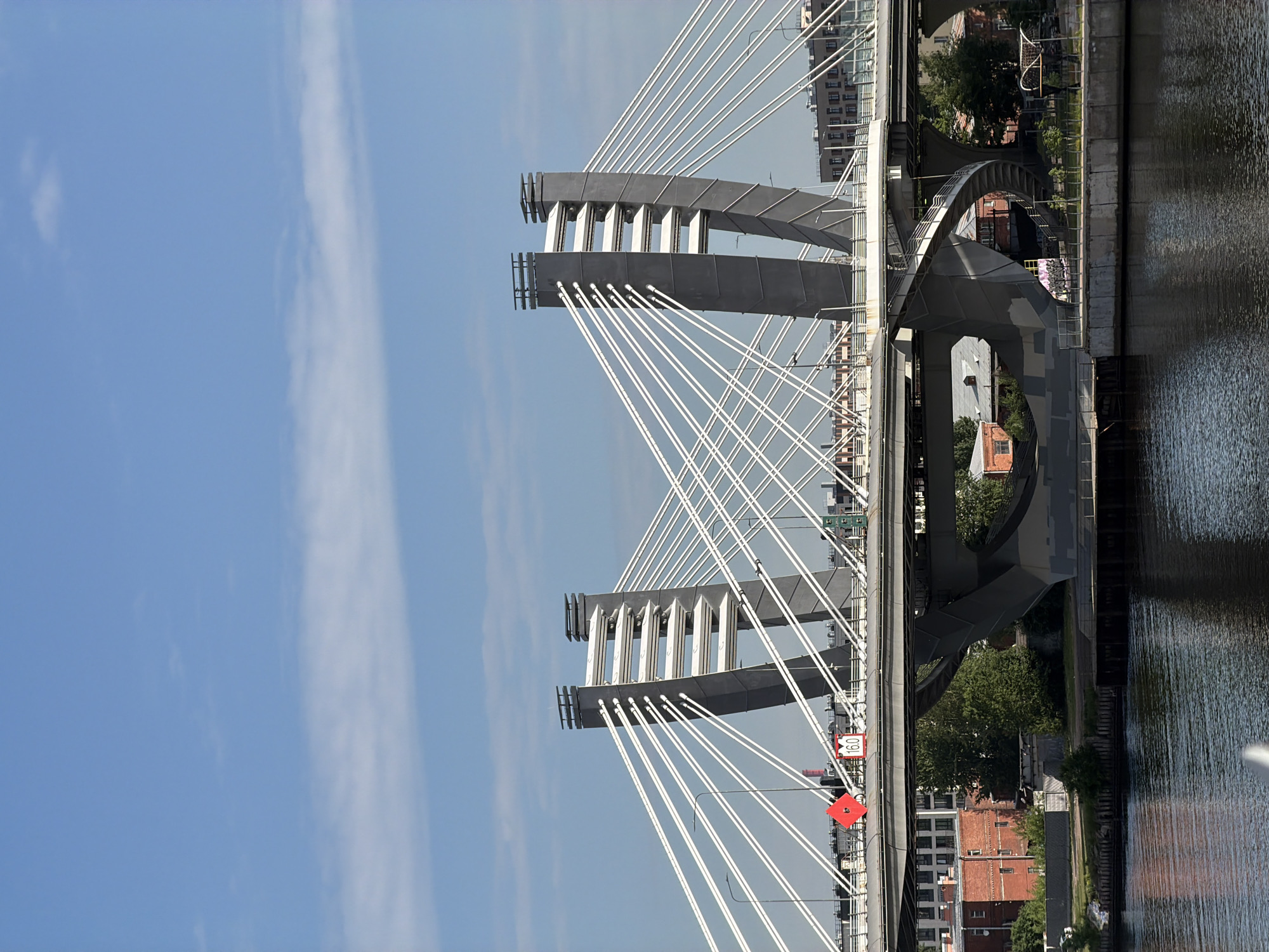 The image shows the cable-stayed Bolshoy Obukhovsky Bridge spanning the Neva River in Saint Petersburg, Russia. Two massive, dark gray towers support the bridge, with numerous white cables fanning out to hold the roadway. The bridge structure is visible, including its complex concrete supports and a spiral staircase leading up to the roadway on the lower level. In the background, buildings with red brick facades and more modern structures are visible along the riverbanks. The sky is blue with scattered white clouds. The water of the Neva River is dark and reflects the sky and bridge. There are no people visible in the image. A red diamond-shaped sign and a sign with "16.0" are attached to the bridge's railing.