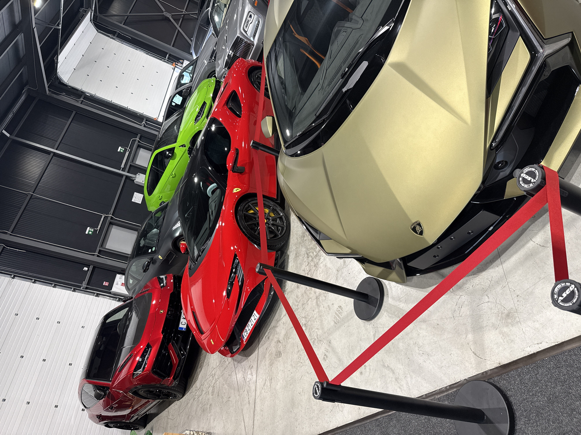The image depicts an indoor car exhibition or showroom located in Plovdiv, Bulgaria. Several luxury vehicles are arranged in a multi-row display, separated from the viewer by a red retractable barrier rope system.
In the immediate foreground, the front section of a matte gold Lamborghini sports car, likely a Huracán or Aventador model, is visible on the right. Its front bumper, hood, and a Lamborghini emblem are discernible.
Behind the barrier, in the middle ground, a red Ferrari SF90 Stradale is prominently featured. Its front fascia, headlights, and yellow brake calipers are visible. The license plate "CB 2121 21" is affixed to its front. To its left, a red Lamborghini Urus SUV with a contrasting black hood and black wheels is positioned. A partial license plate "C KY" is visible on this vehicle. Further to the right and slightly behind the red Urus, a dark matte-colored sports car, likely a Ferrari, is partially obscured.
In the background, a vibrant lime green Lamborghini Urus SUV is visible. To its right, a silver Rolls-Royce Cullinan SUV is parked, recognizable by its distinctive large grille and headlights. Other vehicles are present further in the background but are largely obscured.
The environment features a light gray concrete floor with scuff marks. The walls consist of corrugated metal panels; the left wall is white, while the right wall is black. A large white roll-up garage door is partially open at the top right of the frame. A green emergency exit sign is mounted on the left wall. Black stanchions supporting the red barrier ropes are visible in the foreground. No individuals are present in the image.