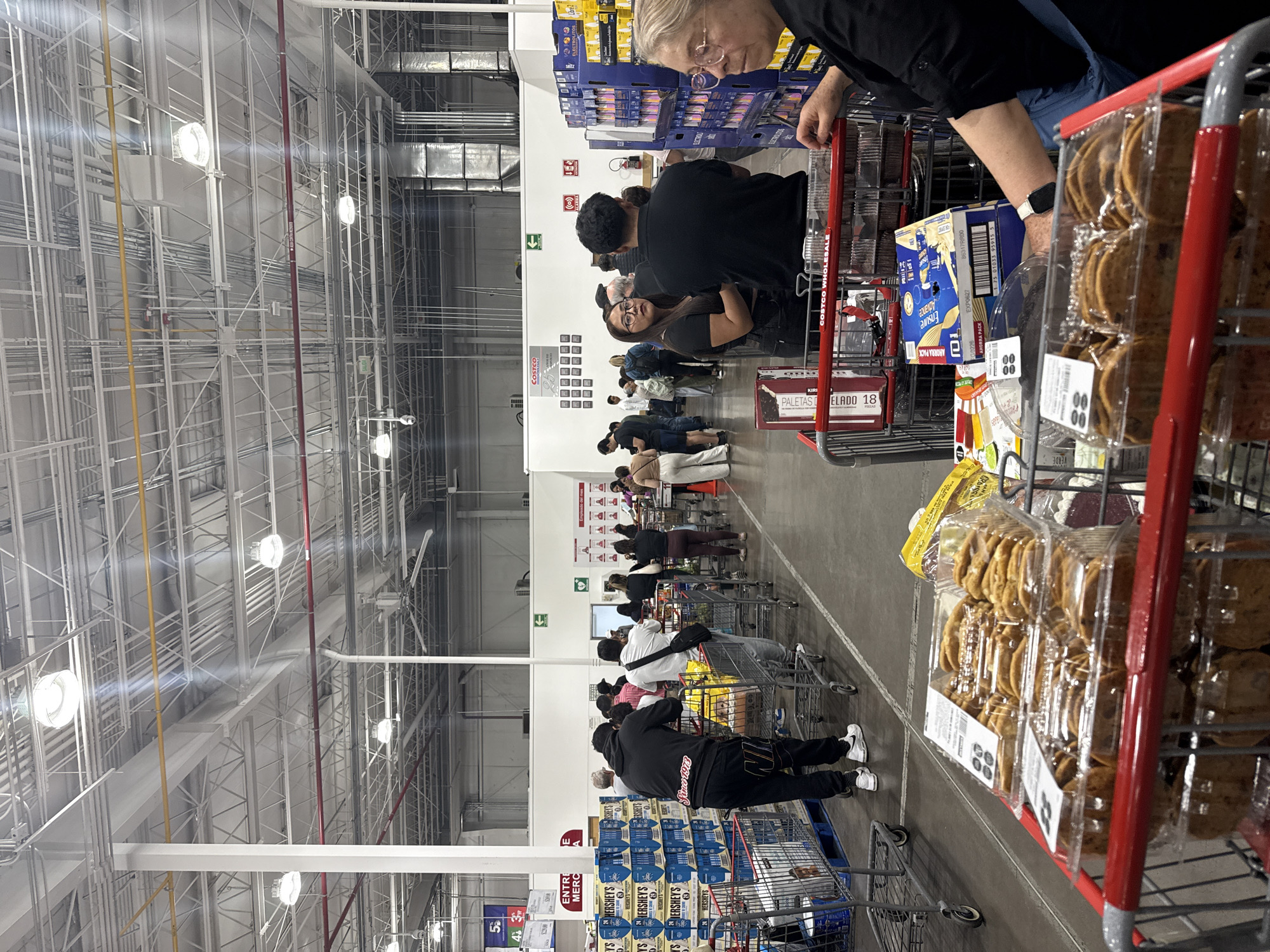 The image shows a long checkout line in a Costco store, likely in Puebla, Mexico. Shoppers with loaded carts are waiting to pay. The carts contain various items, including cookies, bottled water, and other groceries. Several people are in the line, and the store's high ceiling is visible, with overhead lighting and industrial infrastructure. A woman in line looks directly at the camera. Signage in Spanish and English can be seen on the products and store displays. The Costco is filled with several customers and long lines.
