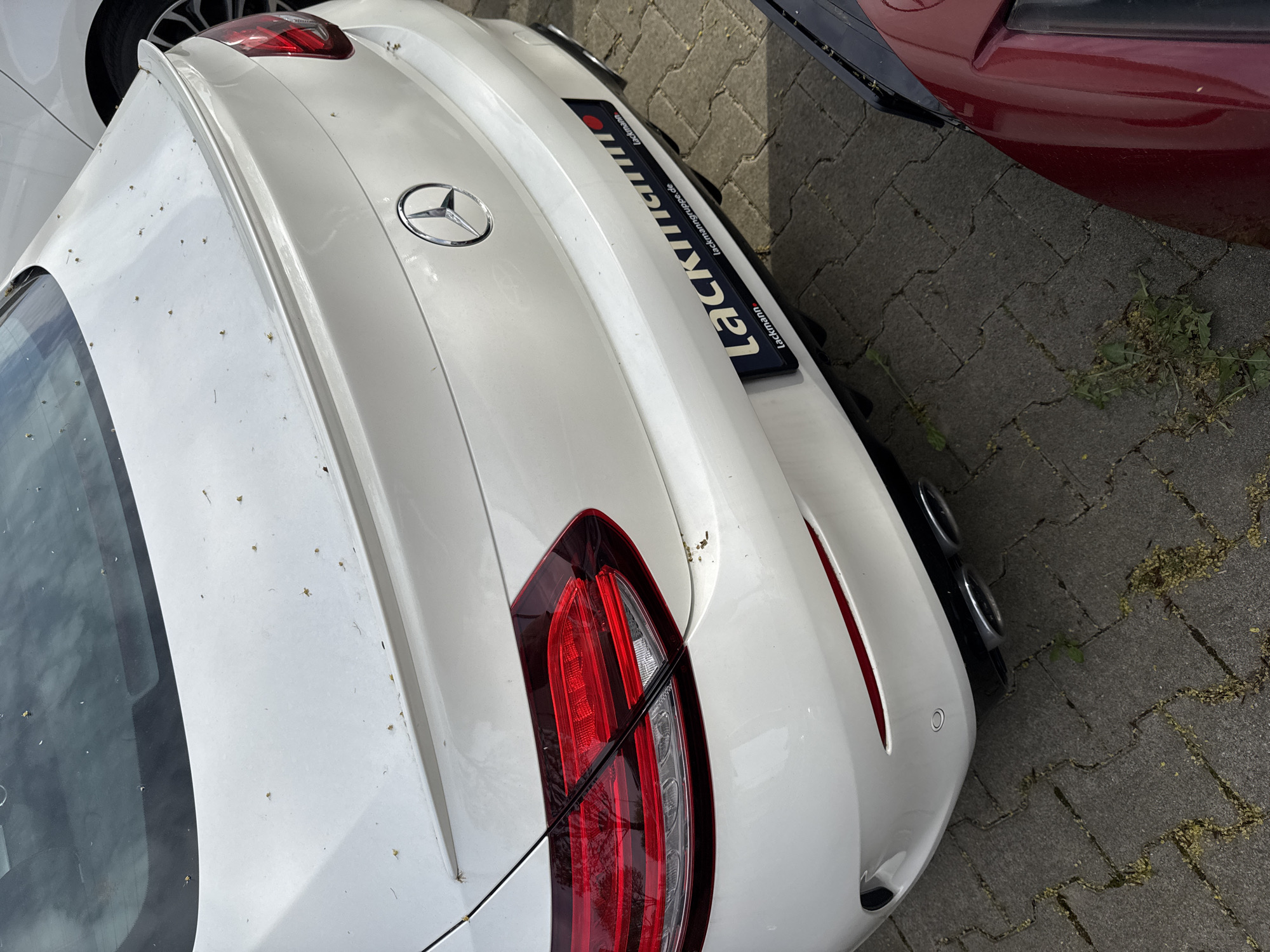 The image shows the rear of a white Mercedes-Benz car parked on a paved surface. The car features a Mercedes-Benz emblem and a license plate frame displaying the name "Lackmann" and the website "lackmanngruppe.de". The car is positioned next to other vehicles, one appearing to be red. There are leaves and small pieces of plant debris scattered on the pavement. Given that Lackmanngruppe has a location in Solingen, Germany, it is plausible that the picture was taken in this city.