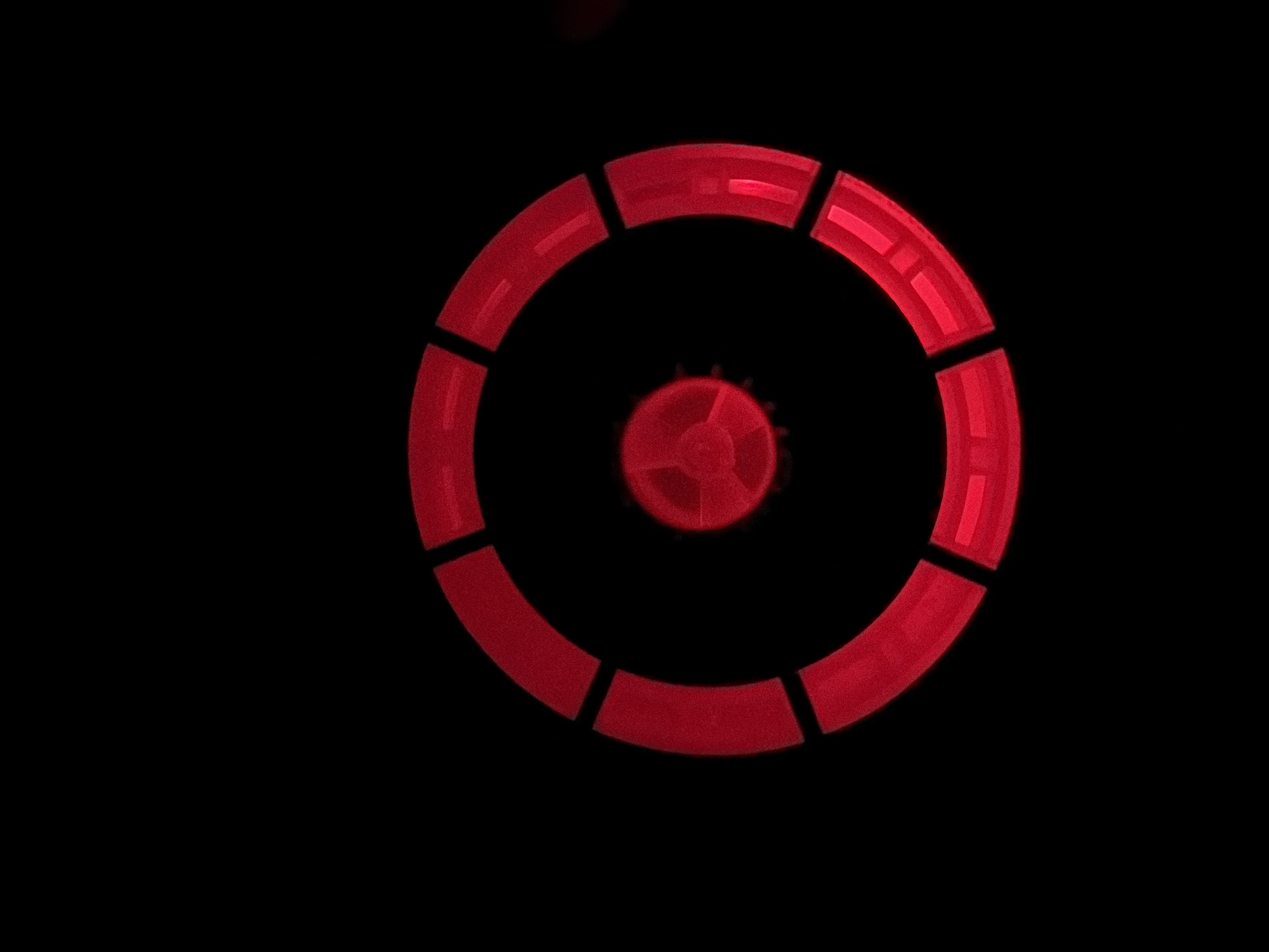 The image is a close-up, overhead shot of a red circular object against a black background. The object appears to be a circular structure composed of several segmented, illuminated red sections arranged around a central, red, fan-like component. The overall impression is that of a technological or mechanical device, possibly a light or a part of a larger machine, set in a dark environment. The location is unknown.