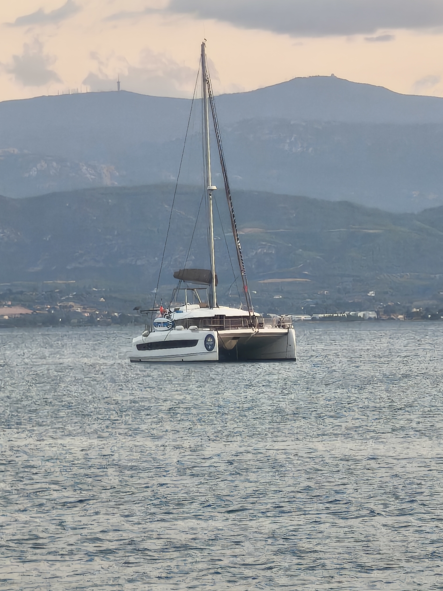 A white catamaran with two hulls is sailing on a calm sea in front of a hazy mountainous landscape. The catamaran has a tall mast with sails furled and rigging. The mountain range in the background is covered in vegetation and appears to have some man-made structures, possibly communication towers, on its peaks. The sky is a soft peach color with some scattered clouds. The overall scene suggests a tranquil day at sea near a coastal area, potentially within the Eretria Municipal Unit of Greece, given the mountainous terrain and the presence of the sea.