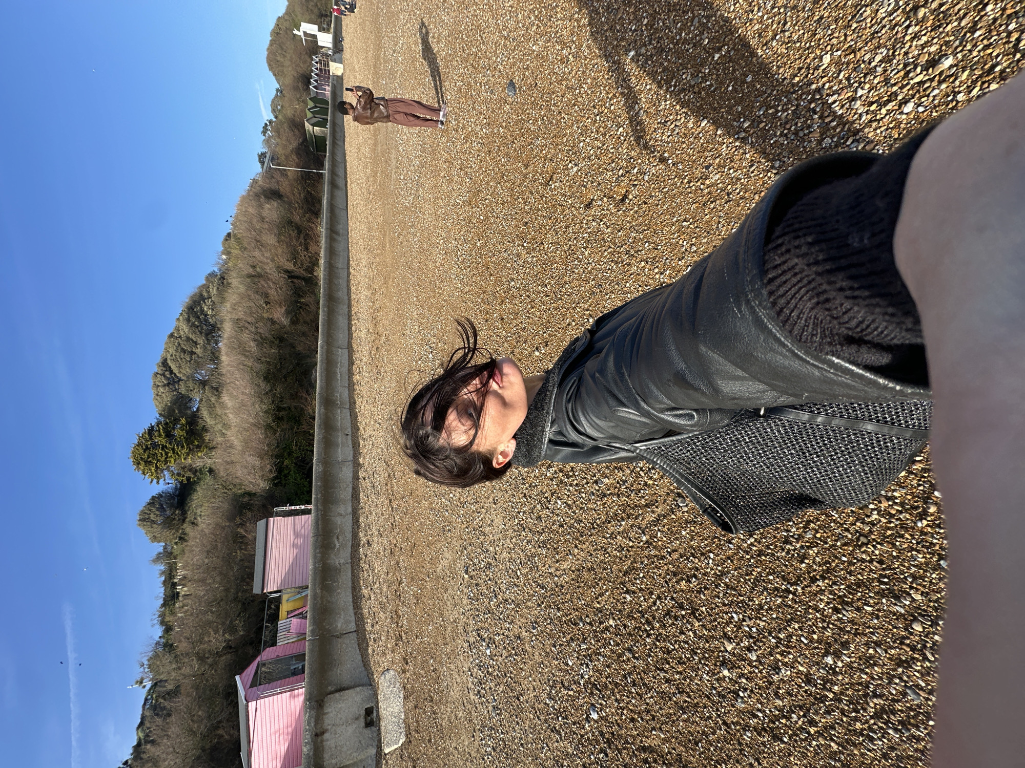 The image shows a shingle beach in Folkestone and Hythe District, United Kingdom, with a woman lying down. She is wearing a black leather jacket and a gray knitted garment, and a patterned bag is visible. In the distance, another person is standing and appearing to take a photo. There are beach huts and vegetation lining the edge of the beach, and the sky is blue.