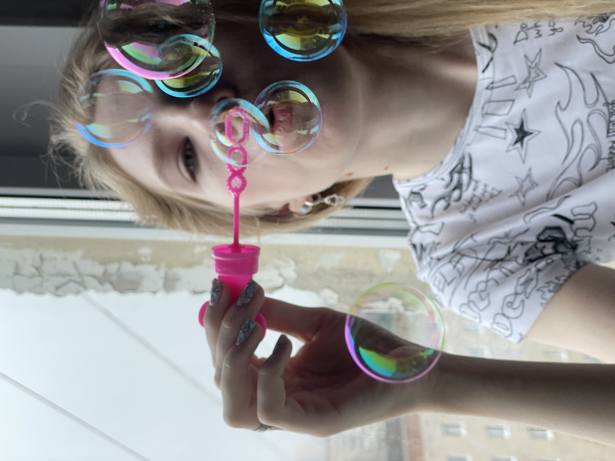 An inverted, eye-level indoor shot captures a person with blonde hair blowing iridescent bubbles near a window in Izhevsk, Russia. The individual, likely female, is depicted from the chest up, with their head positioned to the right of the frame due to the image's 90-degree counter-clockwise rotation. They hold a bright pink bubble wand in their left hand, which features patterned nail polish and a ring on the ring finger. Numerous soap bubbles, reflecting vibrant hues of pink, blue, yellow, and green, float around their face and hand.
The person wears a white t-shirt adorned with black graphic designs, including stars, flames, and stylized text; the phrase "You Will Regret" is legible on the right side of the shirt, along with other symbols like "DMG." Through the window, a muted urban landscape with indistinct building structures is visible, suggesting an overcast daytime. The white window sill and frame are apparent, and the glass appears somewhat obscured. The person maintains a neutral, focused expression while engaging in the activity.