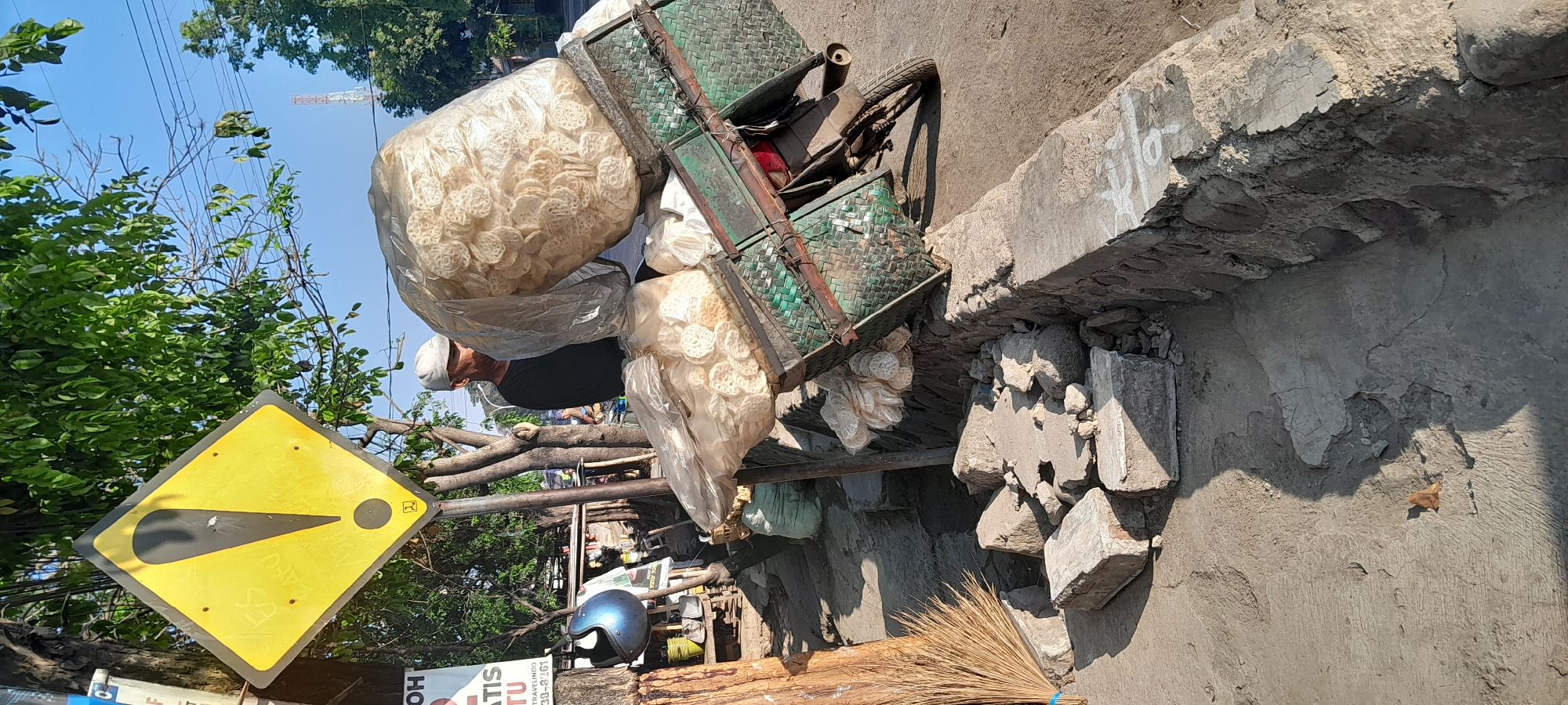 In the city of Tambakrejo, Indonesia, a man wearing a white cap and black shirt pushes a cart laden with large bags of what appear to be fried snacks, possibly kerupuk. The cart is a green woven basket attached to a motorbike. One very large, translucent plastic bag, filled with the snacks, is perched on top of the cart, and a smaller bag is to its left. In the foreground, broken concrete debris is scattered on the dusty ground, next to a stone wall. A broom leans against a wooden post. Behind the man, a yellow diamond-shaped warning sign with a black exclamation mark is visible on a pole, with green foliage and a clear blue sky in the background. A communication tower is visible in the distance.