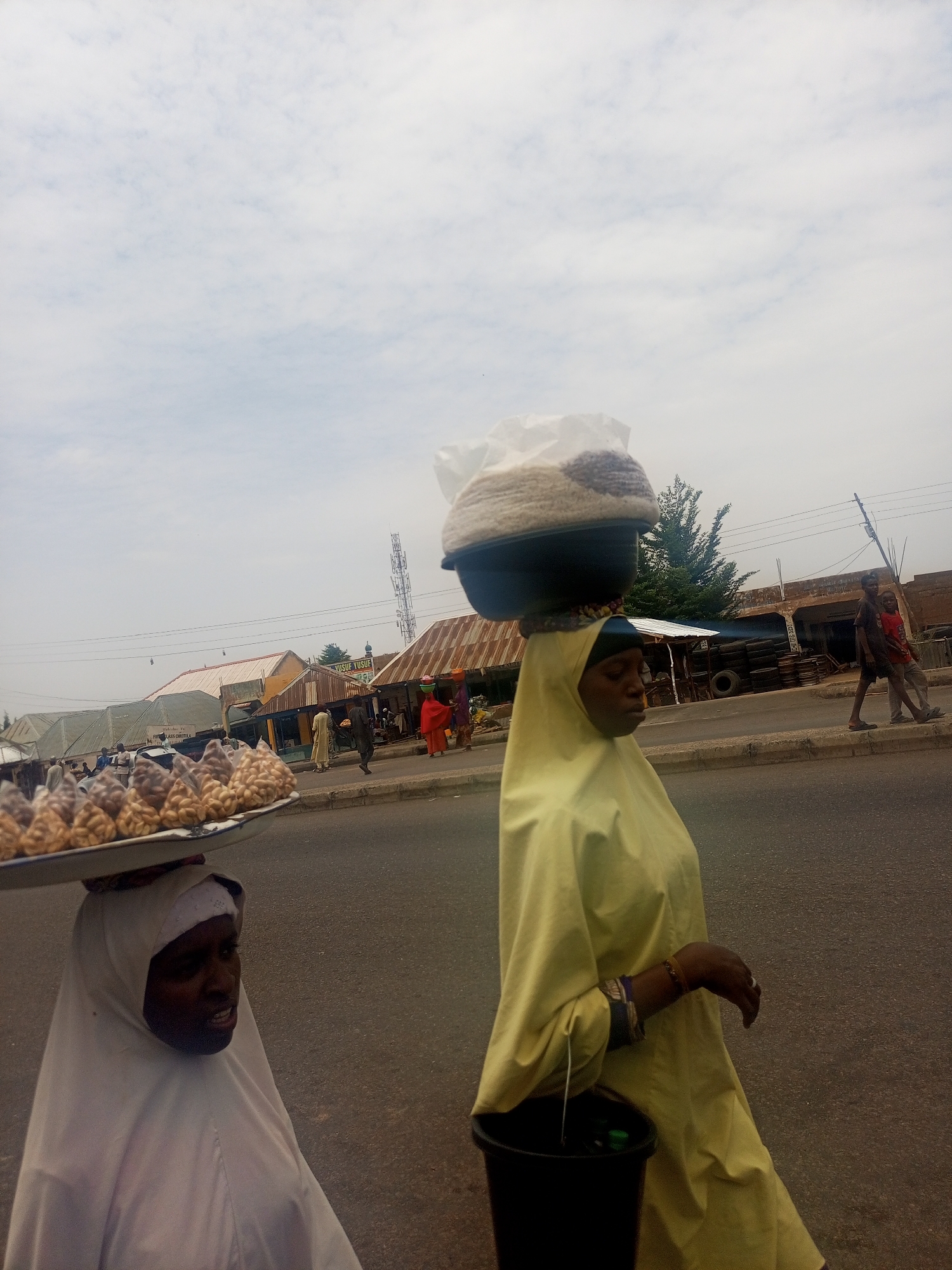 The image captures a street scene in Gombe, Nigeria. Two women are walking along the side of a road. One woman, dressed in white, carries a tray of small bags on her head, likely containing groundnuts. The other woman, wearing a yellow covering, balances a large bowl with a plastic bag on her head and carries a black bucket. In the background, several other people are walking near buildings. One building has the name "YUSUF YUSUF" printed on it. Overhead power lines are visible, as is a cell tower. The road is paved. The sky is overcast.