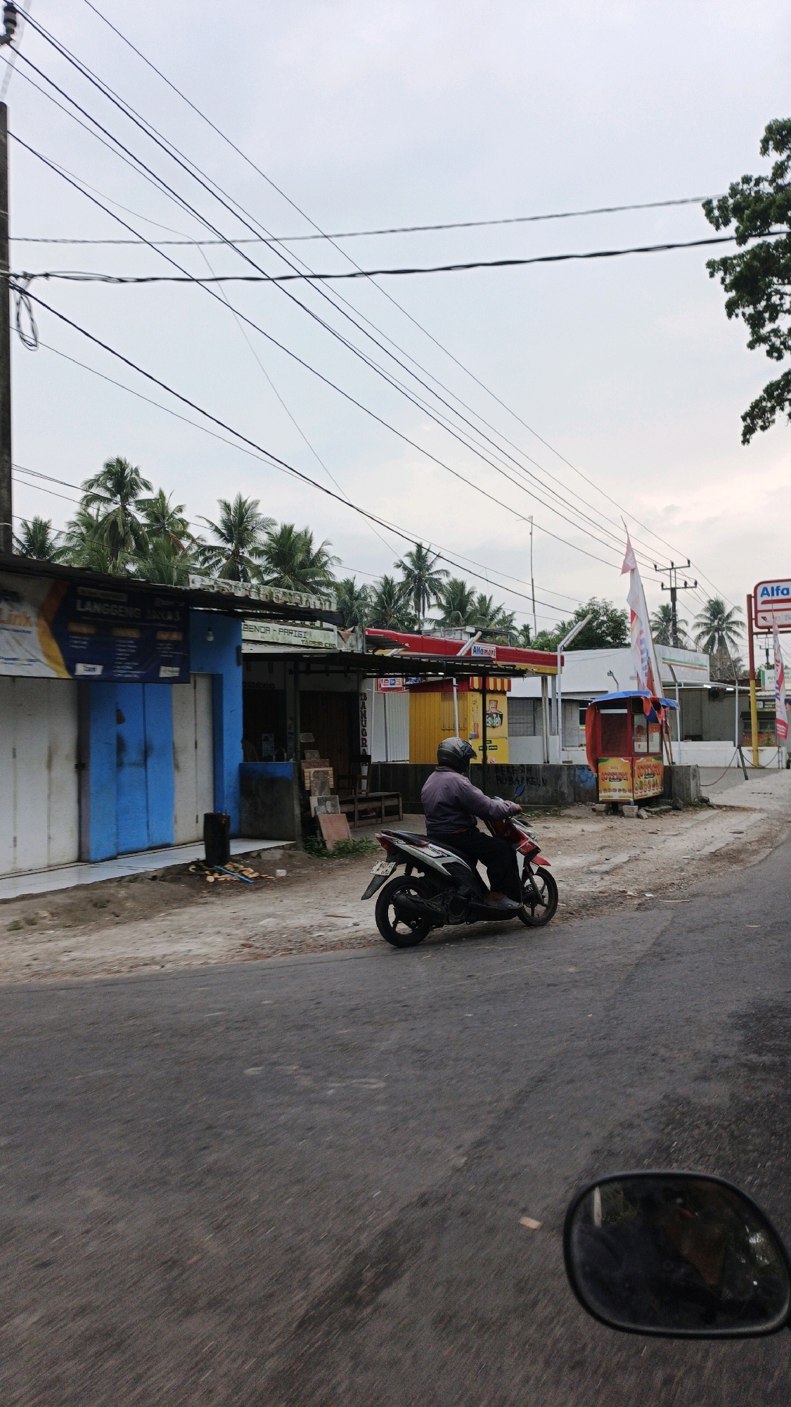 A man in a purple shirt and helmet rides a black and red motorcycle on a paved road. To his left are several buildings with blue and white facades, one with a large blue door. Further down the road, a red and yellow gas station with an Alfamart sign is visible, along with a small red and yellow kiosk. Palm trees line the background under a cloudy sky. The image appears to be taken from another vehicle, with a reflection of the photographer visible in a rearview mirror in the foreground. The location is identified as Cibenda, Indonesia.