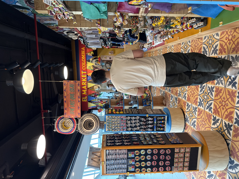 An interior view depicts a souvenir shop, identified by a banner reading "EL MARKET COLOMBIA" and another sign stating "¡PA' LAS QUE SEA!", located within Colombia, specifically inferred to be in the Area Metropolitana Centro Occidente (AMCO). The shop features a black exposed ceiling with red piping and multiple industrial-style pendant lights and track lighting fixtures. The floor is tiled with a distinctive pattern of orange, yellow, red, and black geometric and floral motifs.
A male individual, viewed from behind, occupies the foreground and middle ground, walking away from the camera towards the rear of the store. He wears a light beige short-sleeved t-shirt, dark olive-green cargo pants, and light-colored footwear. His dark hair is short. A blurred figure is partially visible deeper within the shop, to the left of the walking individual.
The shop is extensively stocked with merchandise. On the left, two tall, black and yellow rotating display stands present numerous keychains, magnets, and decorative pins. Hanging from the ceiling are two traditional Colombian "sombrero vueltiao" hats and one white fedora-style hat. To the right, a long, multi-tiered shelving unit extends into the background, filled with various souvenir items including small figures, textile bags, and other crafts. Colorful triangular flags (red, yellow, blue) are strung above the main "EL MARKET COLOMBIA" banner. The left side of the scene reveals a large window or glass partition, indicating an open-plan environment.