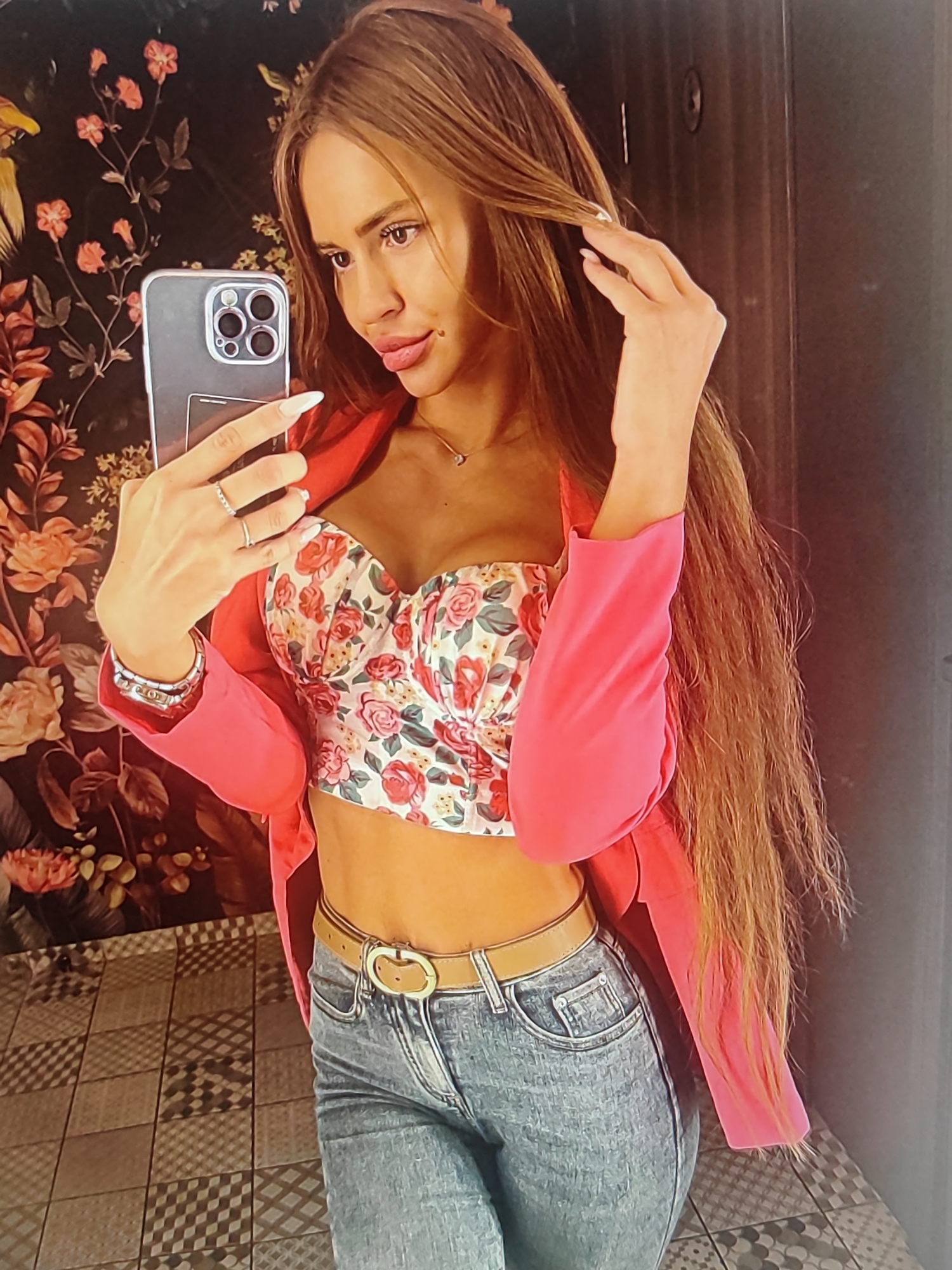 A woman takes a mirror selfie in what appears to be a restroom. She is holding her phone in her left hand, and her right hand is touching her long, brown hair. She is wearing a pink blazer over a floral-patterned, cropped bustier top and blue jeans. A brown belt with a gold buckle cinches her waist. Her fingernails are long and white. The background shows a dark, floral-patterned wallpaper and a tiled floor. The lighting is dim, suggesting it is likely nighttime or indoors. The woman's expression is neutral. There is no visible text in the image. The setting appears to be a stylish, possibly modern, interior space, given the wallpaper and tile choices.