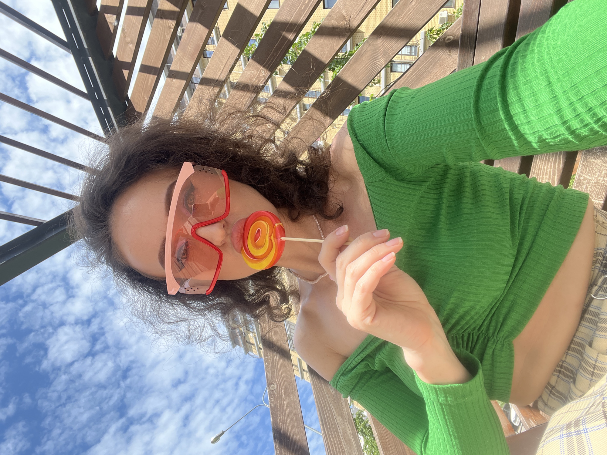 A woman with curly brown hair and pink-tinted sunglasses is seated on a wooden bench outdoors, holding a red and yellow lollipop to her lips. She is wearing an off-the-shoulder green top and plaid pants. The background features a wooden structure overhead, a blue sky with scattered clouds, and a building in the distance. The image was taken at a location identified as Kazan, Russia.