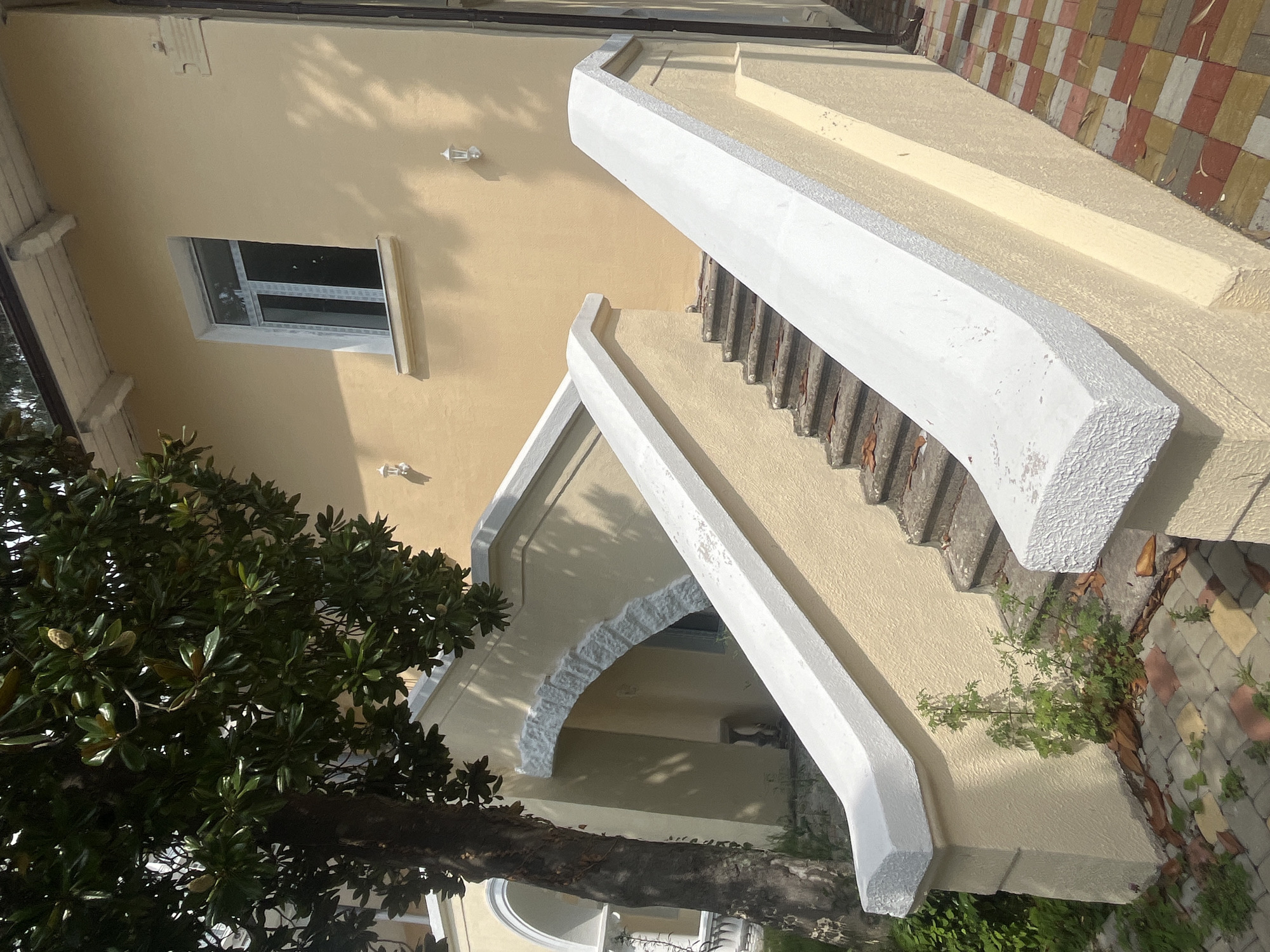 The image shows an exterior view of a light yellow stucco building with a white-trimmed window on the second floor. A large tree with dark bark and dark green leaves stands to the left of the building, partially obscuring an arched entryway. A concrete staircase with white stucco railings ascends from the foreground towards the building. The stairs are weathered and have some debris on them. The ground in front of the stairs is paved with interlocking bricks in red, yellow, and gray. The scene appears to be set in a residential or hotel area, possibly in Sokhumi, Abkhazia, given the architectural style and vegetation. No people are visible in the image.