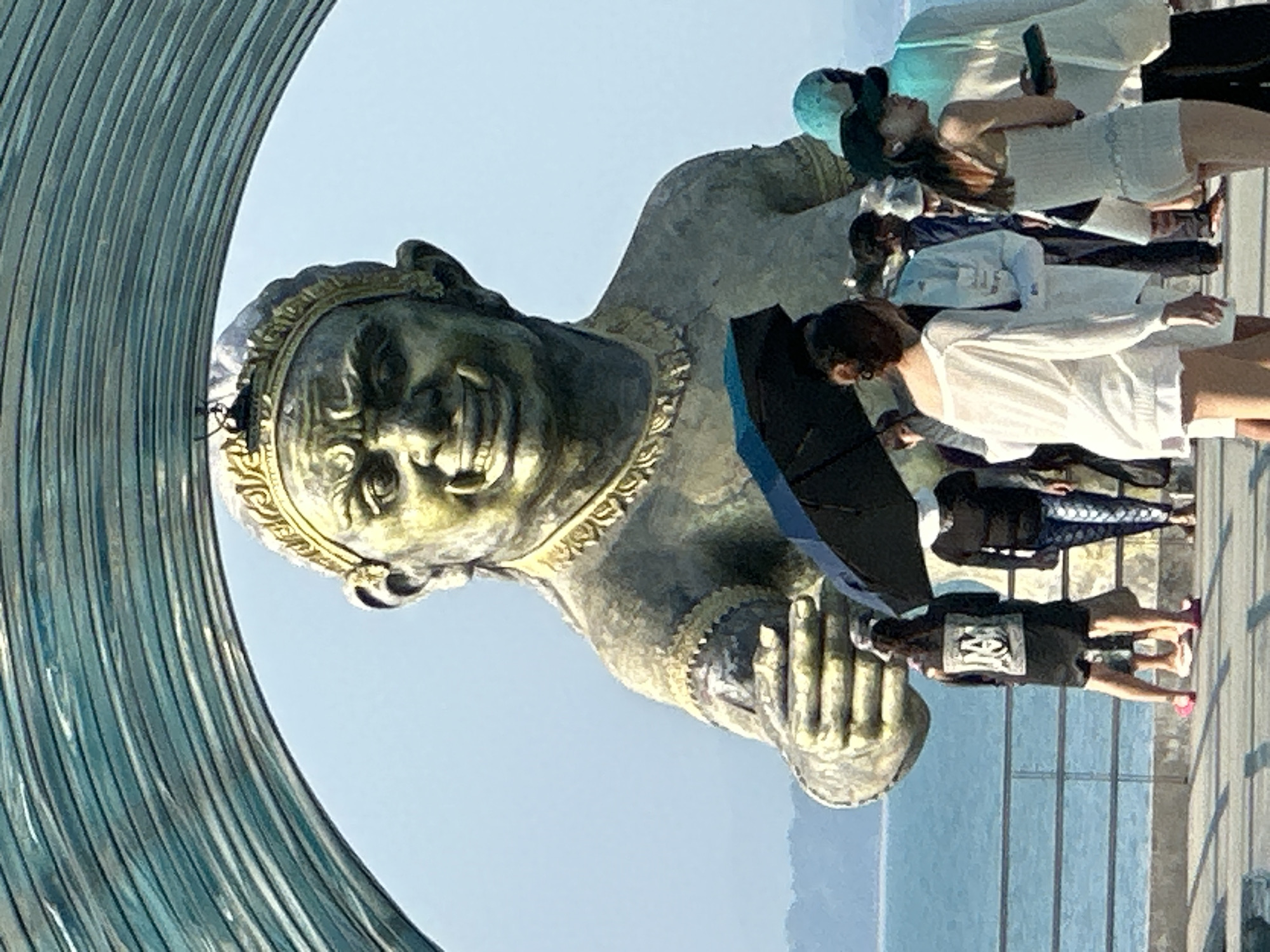 A low-angle, medium shot shows a statue in Ao Lukyon, Thailand, with a group of people in the background. The statue is of a weathered, gold-colored man with a stern expression. He is holding his hands together in a praying gesture. He wears a decorative collar and headdress. To the left of the statue, a section of a modern curved blue metal structure is visible.
In the background, a group of people is standing on a concrete walkway. To the right are a man with an umbrella, woman in white shirt, woman looking up at the statue. Beyond the walkway, the blue sea and a hazy mountain range are visible. The sky is light blue.