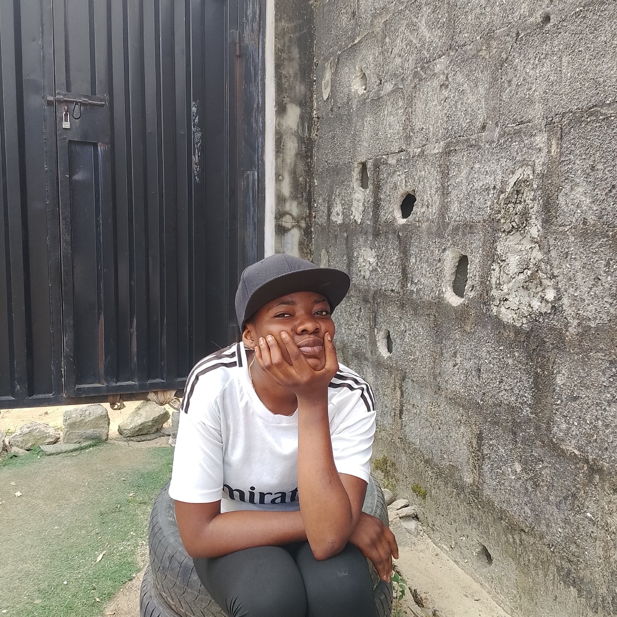In Ado, Nigeria, a young Black person is depicted sitting on a stack of tires. They are wearing a black baseball cap, a white t-shirt with black stripes on the sleeves, and black leggings. Their right hand is placed on their chin, with their fingers extended. The background features a corrugated metal gate on the left and a textured concrete wall with several holes on the right. The ground is a mix of sand, small rocks, and patchy grass.