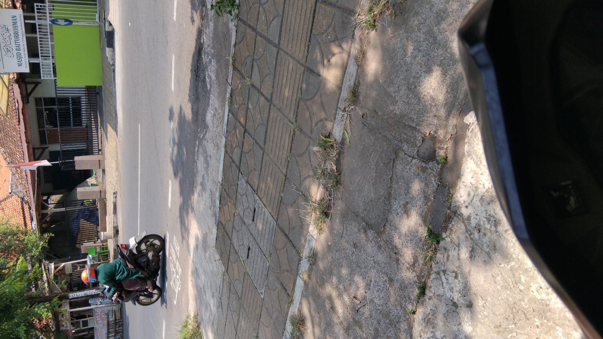 In Proyonangg, Indonesia, a person wearing a green robe and an orange helmet rides a black motorcycle with a white license plate on a paved road. The person is on the right side of the road. To the left of the road, a sidewalk made of grey concrete tiles with some vegetation growing between them is visible. Behind the motorcycle, a building with a red tile roof is partially visible, with a sign that reads "MASJID BAITURROHMAN" in Arabic and English. A green fence is in front of the building. The sky is bright, indicating daylight. The image is taken from a low angle, possibly from another motorcycle.