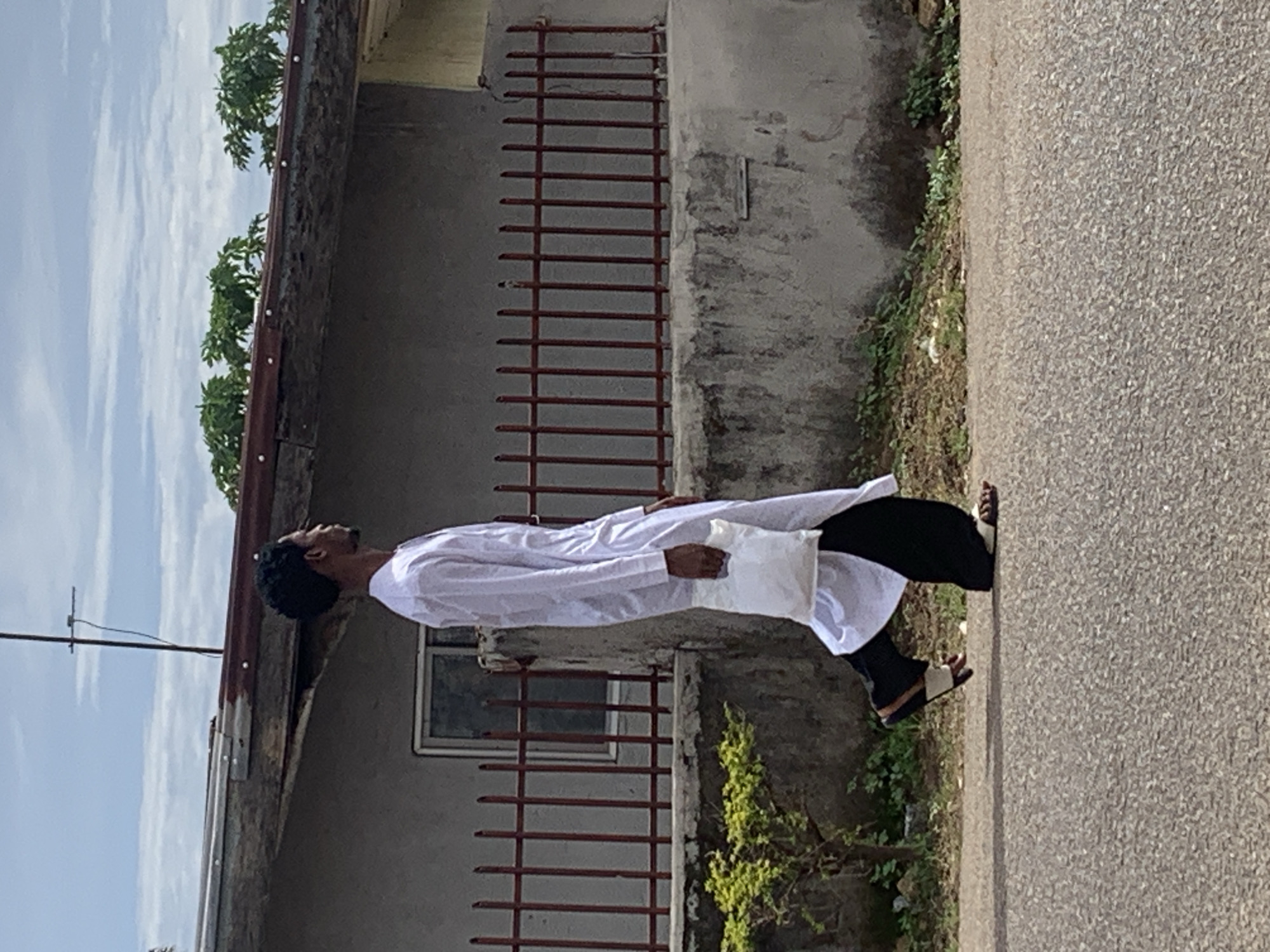 In the city of Jos, Nigeria, an outdoor scene depicts a single adult male walking from left to right on a paved asphalt surface. The man, seen in profile, has dark skin, short curly hair, and a slight beard. He is dressed in a long, loose-fitting white tunic that extends over dark, loose trousers. On his feet, he wears dark-strapped sandals. His right hand holds a white, somewhat crumpled plastic bag, which hangs at his side.
In the background, a single-story building with a light grey wall is visible. A rectangular window with a light frame is set into this wall. The building features a low, red metal fence with vertical bars mounted on a concrete wall section. Above the main wall, a reddish-brown corrugated metal roof is present, from which a vertical antenna extends into the cloudy sky. Small clusters of green foliage are visible above the roofline. The immediate ground between the paved surface and the building wall contains sparse vegetation and earth. The sky is overcast with light grey and white clouds.