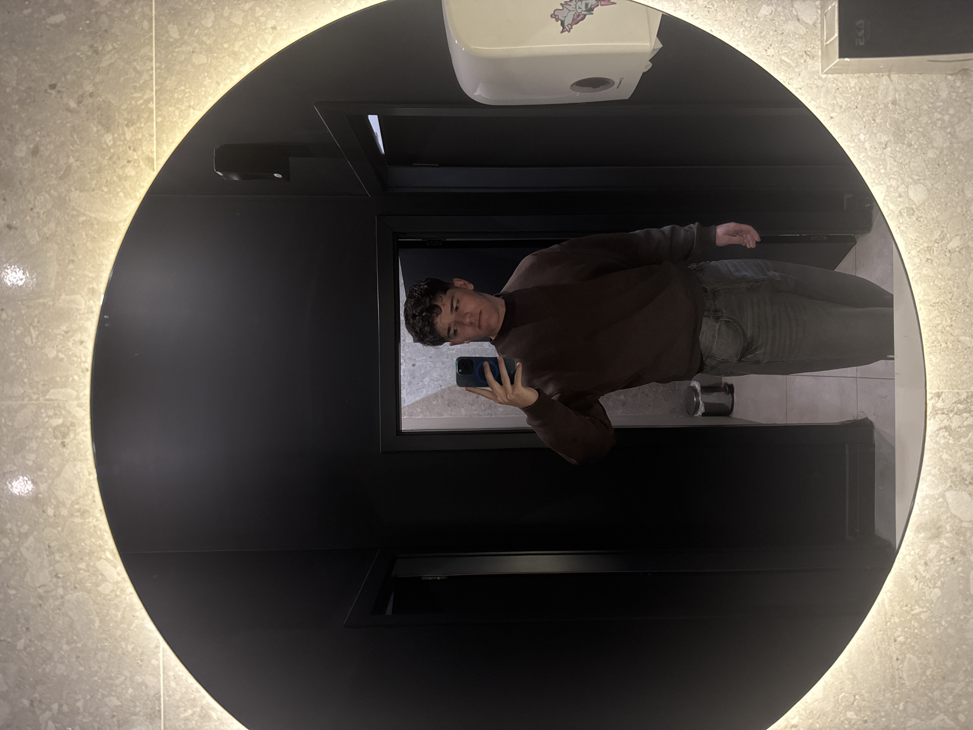 A medium shot captured in a bathroom, reflected in a large circular mirror with an illuminated edge. A person is standing in front of the mirror, holding a blue mobile phone, taking a selfie. The person has short, curly dark hair and is wearing a brown sweater and faded grey jeans. Behind the person is a dimly lit bathroom interior with dark fixtures. Above the person in the reflection are bathroom amenities such as a soap dispenser and a box, seemingly mounted on the wall. The bathroom walls are clad in light-colored stone or tile. This image could have been taken in any bathroom with these characteristics; without additional context, pinpointing the exact location in Sofia, Bulgaria, is not possible.