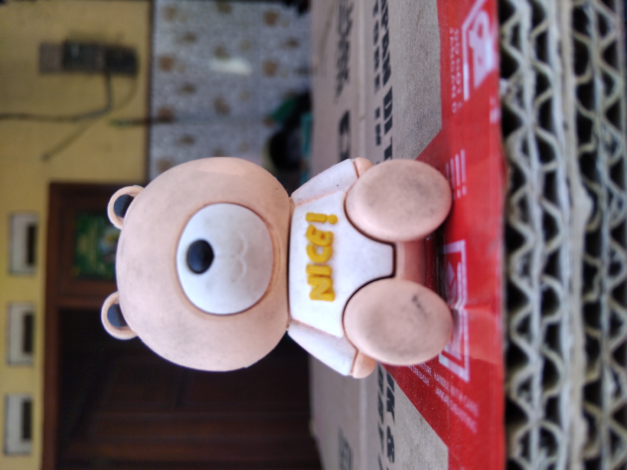 A close-up, low-angle shot shows a small, tan-colored bear figurine sitting on a red and white striped surface, which is itself resting on a stack of corrugated cardboard. The bear wears a white shirt with the word "NICE!" in yellow letters. The figurine's eyes are black, and its nose is black and oval. The background is blurred, but suggests an outdoor or semi-outdoor setting with a yellow wall and a dark doorway. The scene is likely within the city of Kepuh Kiriman, Indonesia, indicated by the context provided.