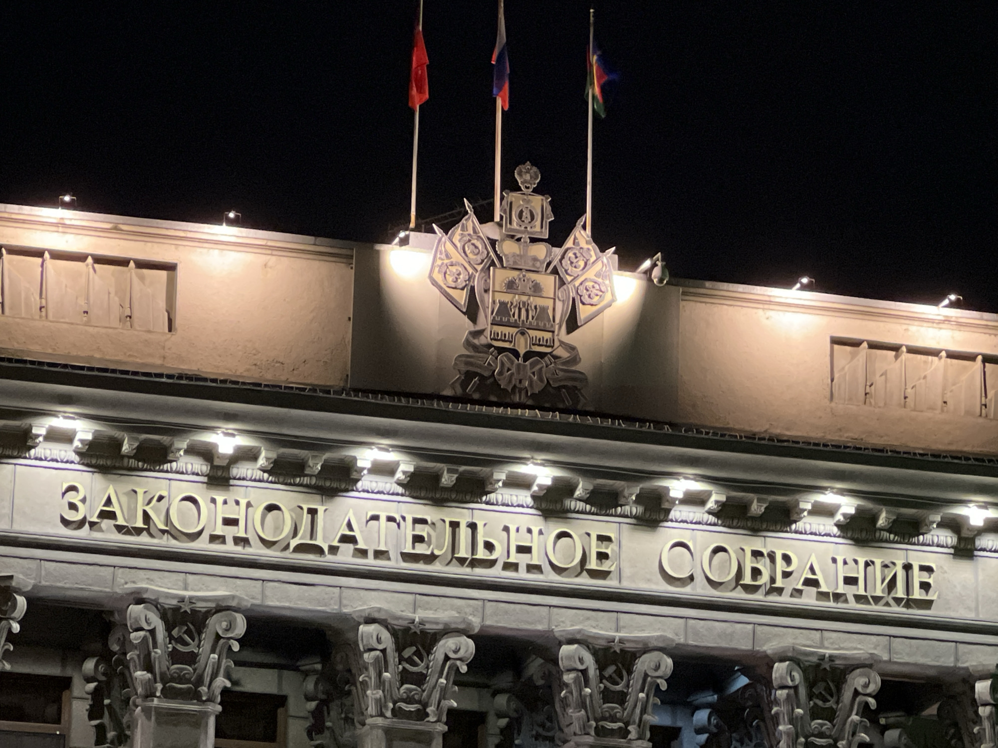 The image shows the front of the Legislative Assembly (Законодательное собрание) building in Krasnodar, Russia at night. The building's facade is lit up, highlighting the inscription "ЗАКОНОДАТЕЛЬНОЕ СОБРАНИЕ" in Cyrillic. Above the inscription is the Coat of arms of Krasnodar krai and three flag poles displaying the Russian flag, Krasnodar krai flag, and unknown red flag. Columns with the symbol of the Soviet Union (hammer and sickle) are visible beneath the inscription.