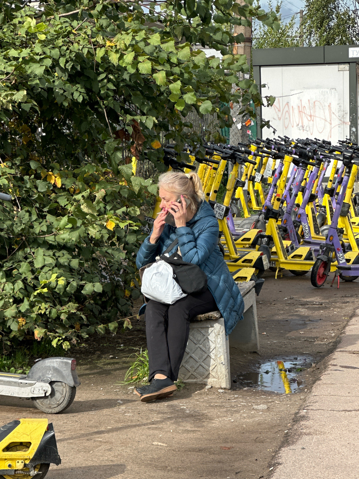 In Vsevolozhsk, Russia, a middle-aged woman with blonde hair pulled into a ponytail sits on a concrete bench near a row of parked electric scooters. She is wearing a dark blue quilted jacket, black pants, and dark shoes. The woman is holding a cigarette in her right hand and talking on her cell phone, which is held to her left ear. A large white bag rests on her lap, and a black bag is slung over her shoulder. She is positioned beneath a large tree with green leaves, some of which are beginning to turn yellow, suggesting an autumn season. To her right, a large number of yellow and purple electric scooters are neatly parked in rows. Behind the scooters, a white structure with graffiti on it is visible. In the foreground, two more electric scooters are parked on the pavement. A puddle of water on the ground reflects the yellow of the scooters. The overall scene depicts an urban outdoor setting with elements of transportation and nature.