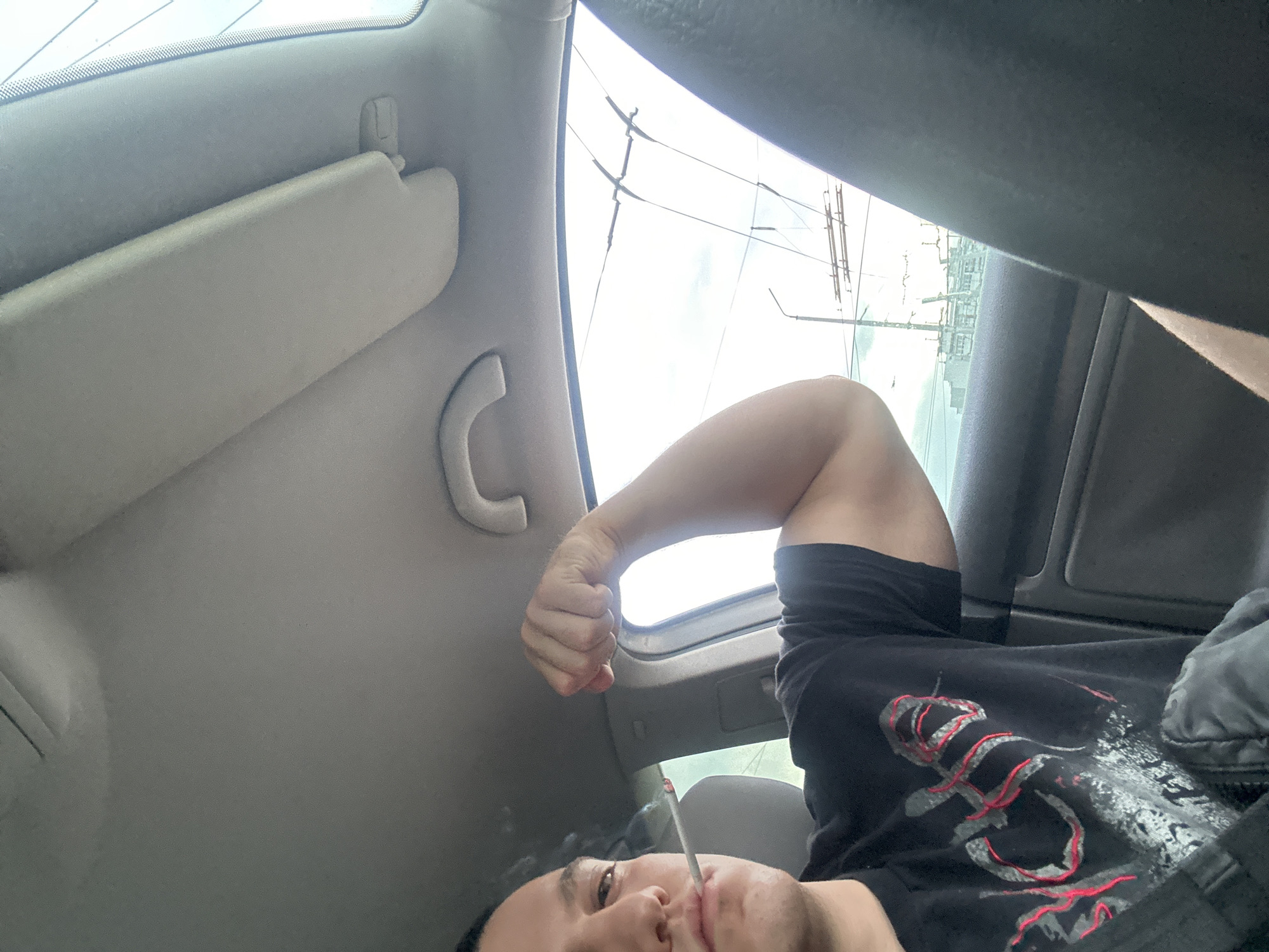 A person is lying inside a car, smoking. The car's interior includes a light gray ceiling, a sun visor, and a grab handle. Outside the car window, power lines and buildings are visible. It's difficult to discern the specific location in Burgas, Bulgaria, from this image.