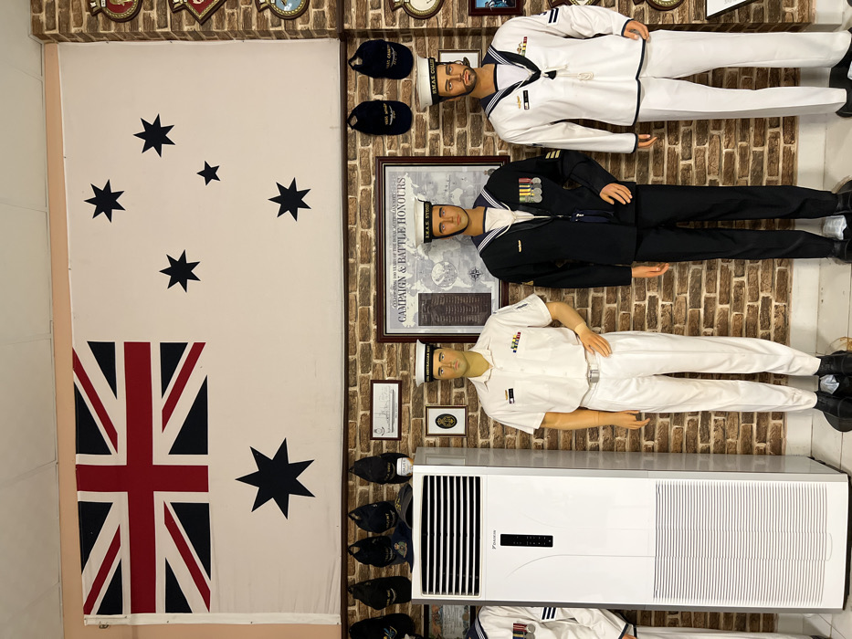 The image displays a room showcasing Australian naval history. A large Australian flag hangs on the left wall, decorated with stars. Three mannequins dressed in navy uniforms stand on the right, representing different ranks. Behind the middle figure, a framed "Campaign & Battle Honours" poster is visible. An air conditioner is placed in the bottom right corner, and naval caps are displayed along the wall. Additional naval decorations are present on the upper wall, and historical documents are framed on the wall above the air conditioner.