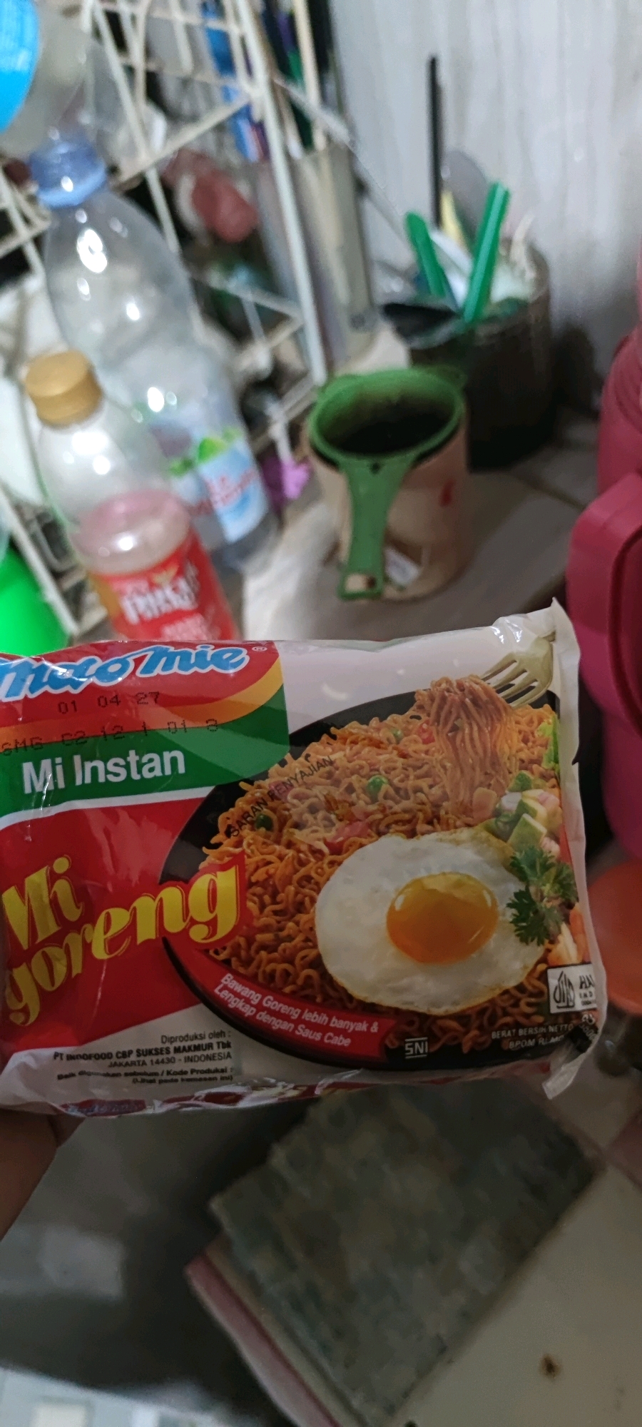 A close-up shot shows a package of Indomie Mi Goreng instant noodles, with a fried egg on top, being held by a hand. The packaging is primarily red and green, with text in white and yellow. The noodles are a reddish-brown color, mixed with vegetables and topped with a fried egg with a bright orange yolk. The background is blurred and appears to be an indoor setting, possibly a kitchen or storage area, with various containers and bottles visible. The scene appears to be in Gambarsari, Indonesia, as indicated by the text on the packaging.