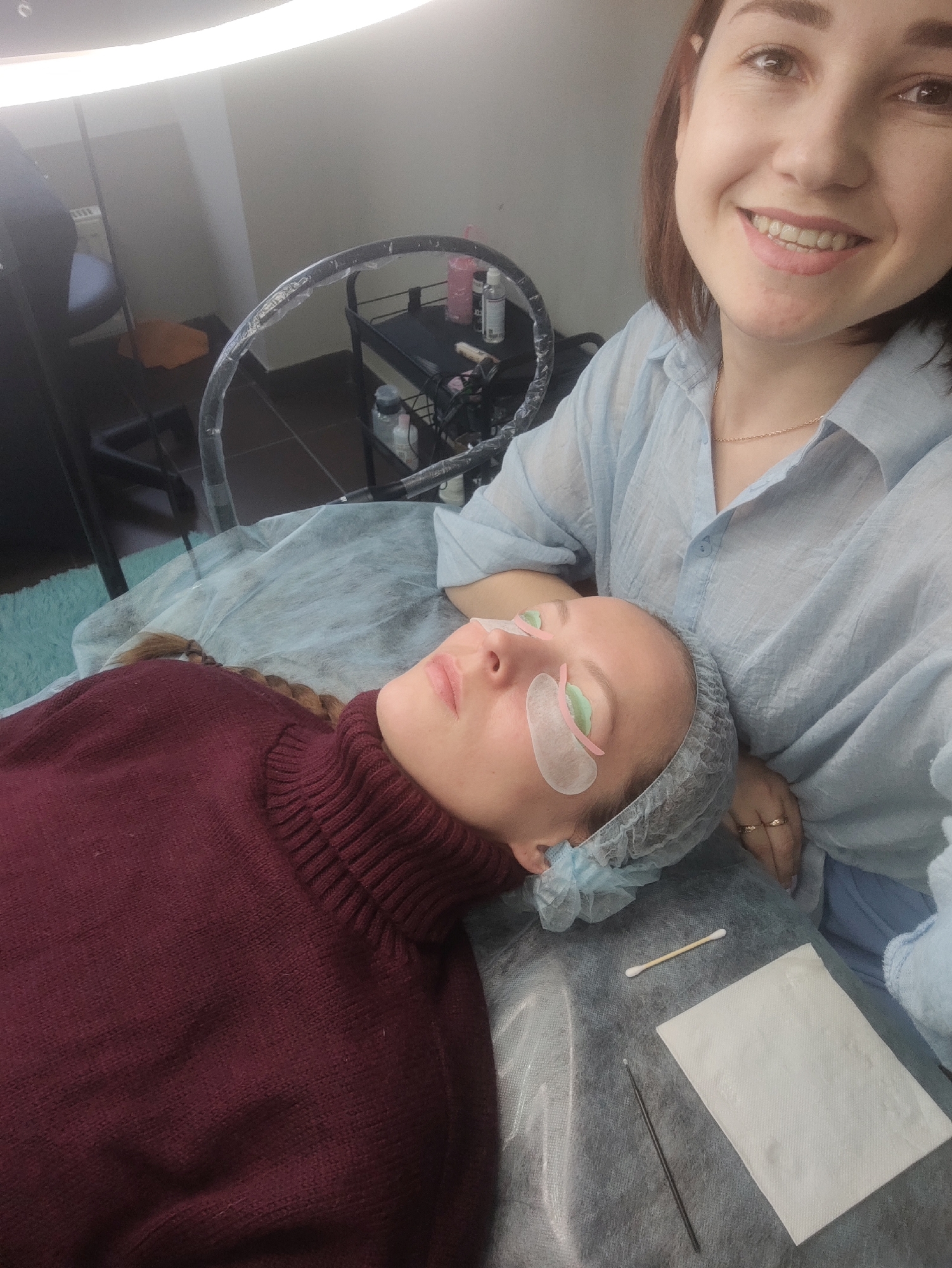 The image depicts a woman lying on a treatment table with eye patches, presumably receiving eyelash extensions or a similar cosmetic procedure. She is wearing a hair net and a maroon turtleneck sweater. A second woman, presumably the technician, is standing beside her, smiling at the camera. She is wearing a light blue button-down shirt and a gold necklace. The scene is illuminated by a ring light above the treatment table. A metal rolling cart with various bottles and equipment is visible in the background. On the treatment table are a cotton swab, a folded paper towel, and a thin metal tool. The city of Chaikovsky, Russia, is not explicitly identified in the image and cannot be definitively determined from the visual information.