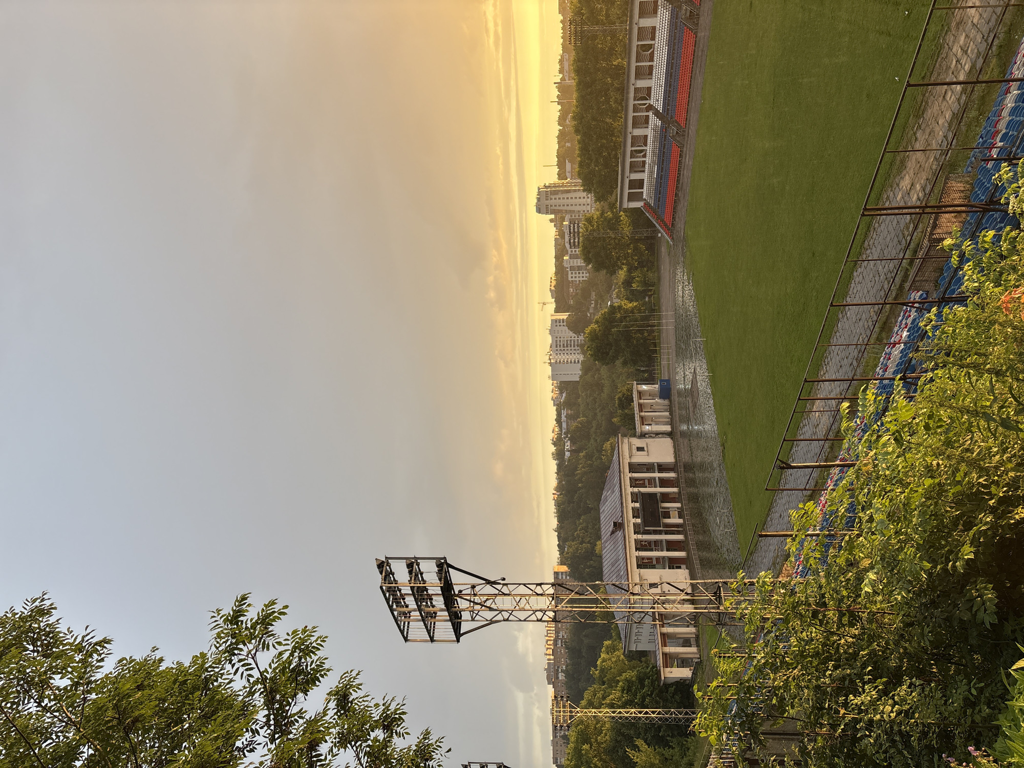 This image depicts an outdoor sports stadium at sunset in Sof'ino, Russia. The sky is a warm orange and yellow, with scattered clouds. A lush green grass field occupies the center of the frame, with empty stadium seating in the foreground and along the far right side. The seating is a mix of blue, red, and white chairs.
In the middle ground, there is a long, white building with columns and an arched entrance. Adjacent to this building is a tall, metal floodlight tower. The area around the building and the edge of the field appears to be wet, suggesting recent rain. Beyond the stadium, a dense line of green trees separates it from a cityscape with several multi-story buildings.
The foreground is dominated by vibrant green foliage and bushes, some of which are illuminated by the setting sun. The overall impression is one of a quiet, slightly damp, and serene evening at a somewhat dated sports facility. There are no people visible in the image.