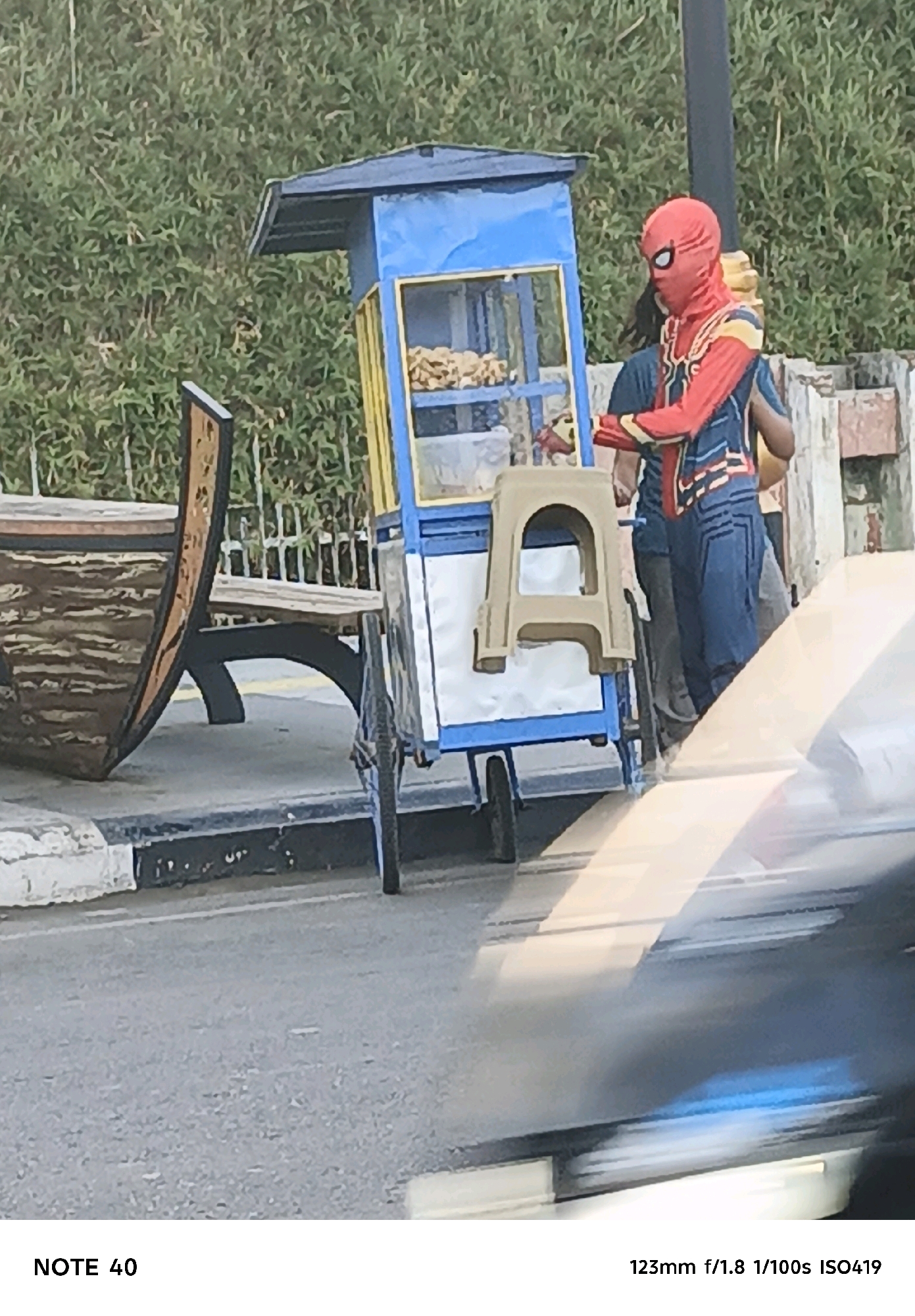 The image depicts a person in a Spider-Man costume standing next to a food cart. The food cart is blue and yellow and contains food items. A plastic stool is attached to the cart. In the background, there is lush green foliage. To the left of the food cart, a wooden bench is visible. The scene appears to be outdoors, possibly on a street or sidewalk in Cilacap, Indonesia, indicated by the text "NOTE 40" and camera settings "123mm f/1.8 1/100s ISO419". The foreground shows a blurred out section of a vehicle.