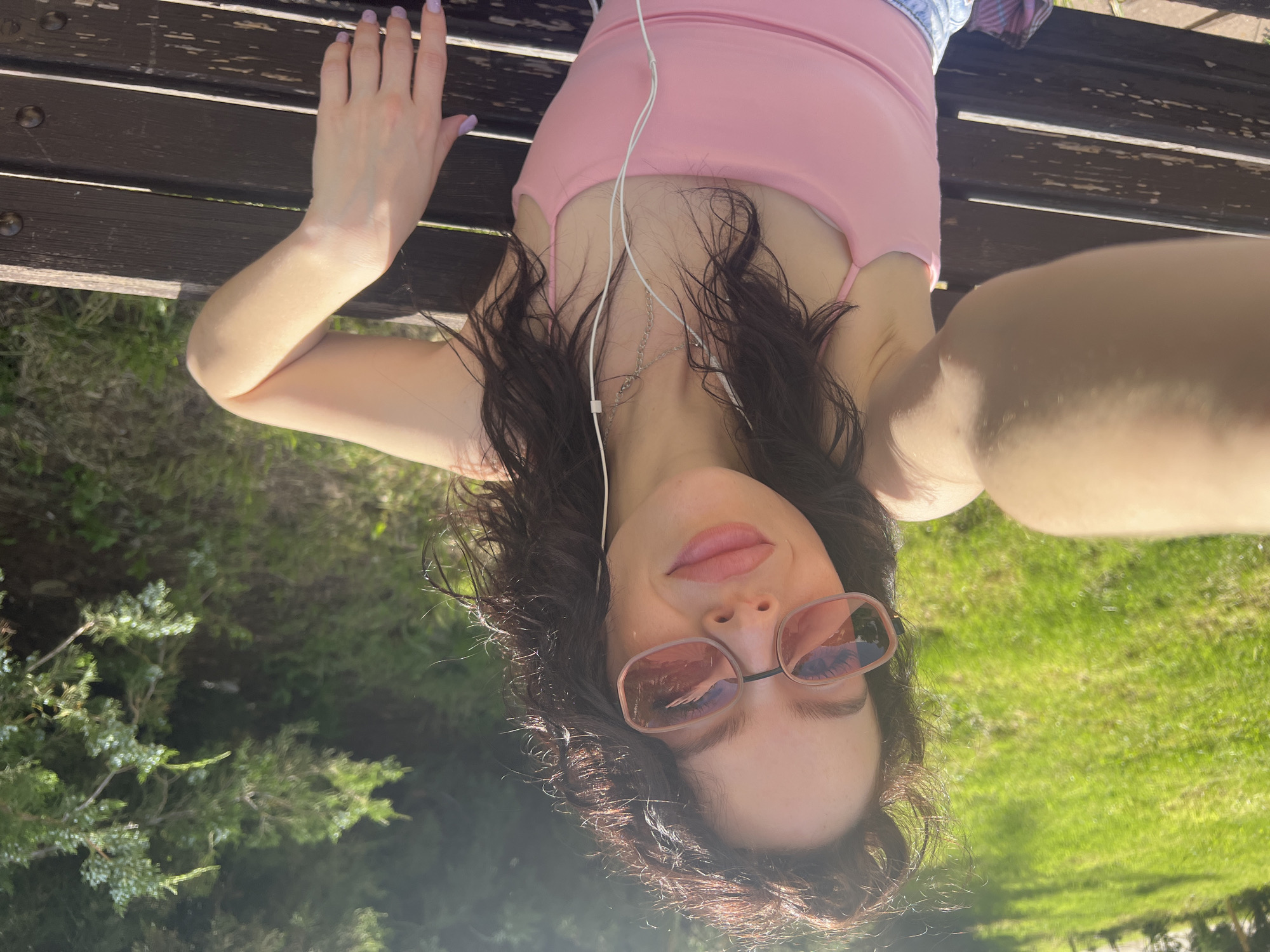 A young woman lies on a dark brown wooden park bench. She is wearing a pink tank top and light-colored shorts. She has headphones in and is wearing pink sunglasses. The background shows green grass and trees. The image was taken in a park. Based on the vegetation and general appearance, the location could potentially be in Krasnodar, Russia.