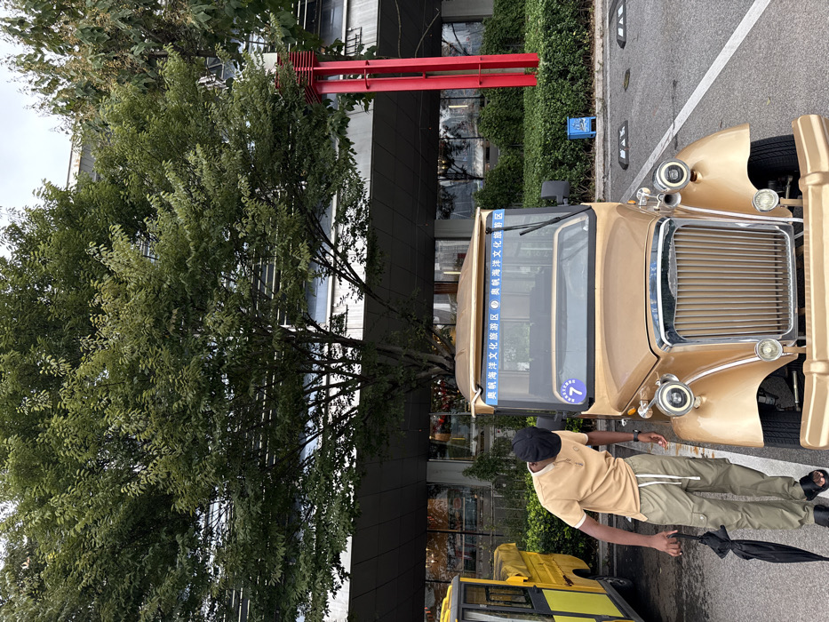 The image captures an outdoor scene in the Olympic Sailing Marine Culture Tourist Area, Shinan District, Qingdao, China. A male individual, appearing to be of Black ethnicity, stands in the foreground, positioned to the left of a gold-colored, vintage-style electric tourist vehicle. He wears a beige short-sleeved polo shirt, olive-green cargo pants with white drawstrings, black slide sandals, and a black flat-brimmed cap. He holds a folded black umbrella loosely in his left hand, and his head is tilted downwards, directed towards the front of the gold vehicle.
The gold vehicle is a four-wheeled, open-top electric tourist transport, featuring a chrome grille, circular headlights, and a windshield banner inscribed with "奥帆海洋文化旅游区" (Olympic Sailing Marine Culture Tourist Area). A blue circular sign displaying the number "7" is affixed below the windshield on the driver's side. Partially visible on the far left, behind the man, is a portion of a yellow utility or golf-cart style vehicle with a black soft top.
The background features a large deciduous tree with dense green foliage, obscuring part of a modern, multi-story building characterized by dark grey paneling and reflective windows. To the right of the gold vehicle, a patch of green shrubs precedes a tall, slender red architectural structure. The ground consists of asphalt pavement marked with white lines, and black curb stoppers are visible on the right. A small blue sign with a white ship emblem is positioned near the curb stoppers.