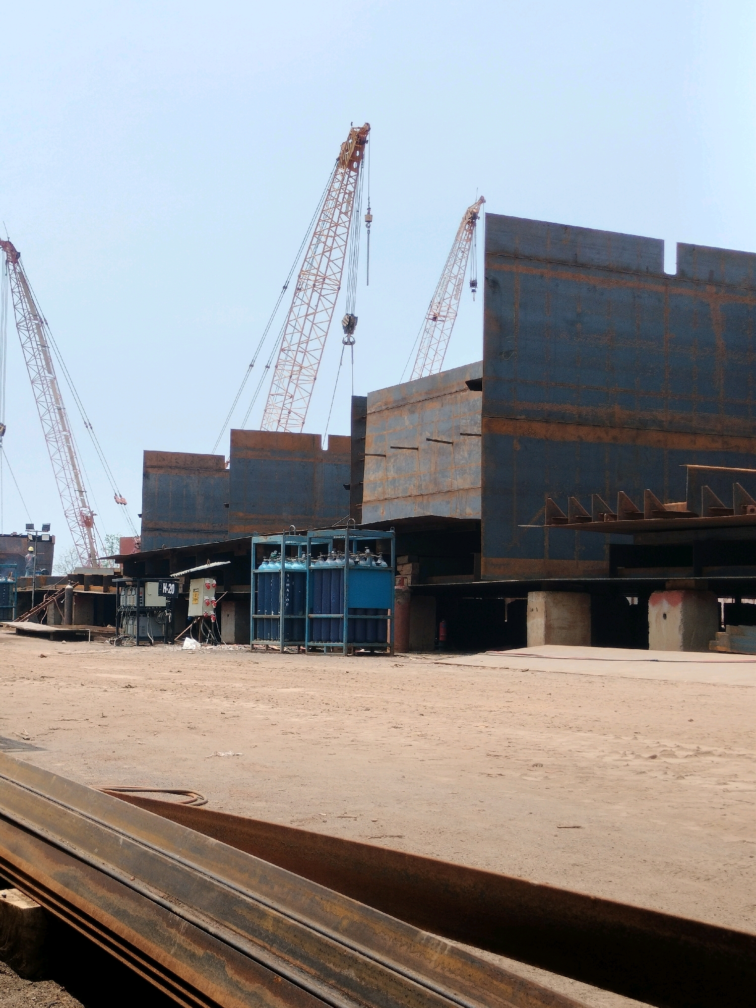 This image depicts a shipyard in Batam, Indonesia, during daylight hours. The scene is dominated by the construction of large metal structures, likely ship hulls or sections thereof, which are dark blue-grey with streaks of rust. Several towering cranes with yellow lattice booms are visible, positioned to lift and assemble these massive components.
In the foreground, piles of long, rusty metal beams lie on the dusty ground. Mid-ground features a collection of blue metal racks holding numerous compressed gas cylinders, suggesting welding or cutting operations are taking place. Electrical equipment, including a distribution box labeled "H-20" and other control panels, are also present. A single worker, wearing a yellow hard hat and a blue shirt, is visible near the electrical equipment.
The environment is an open, industrial area with a dirt or sand floor. The sky is clear and light blue, indicating fair weather. The overall impression is one of ongoing heavy industrial activity within a shipbuilding or repair facility.