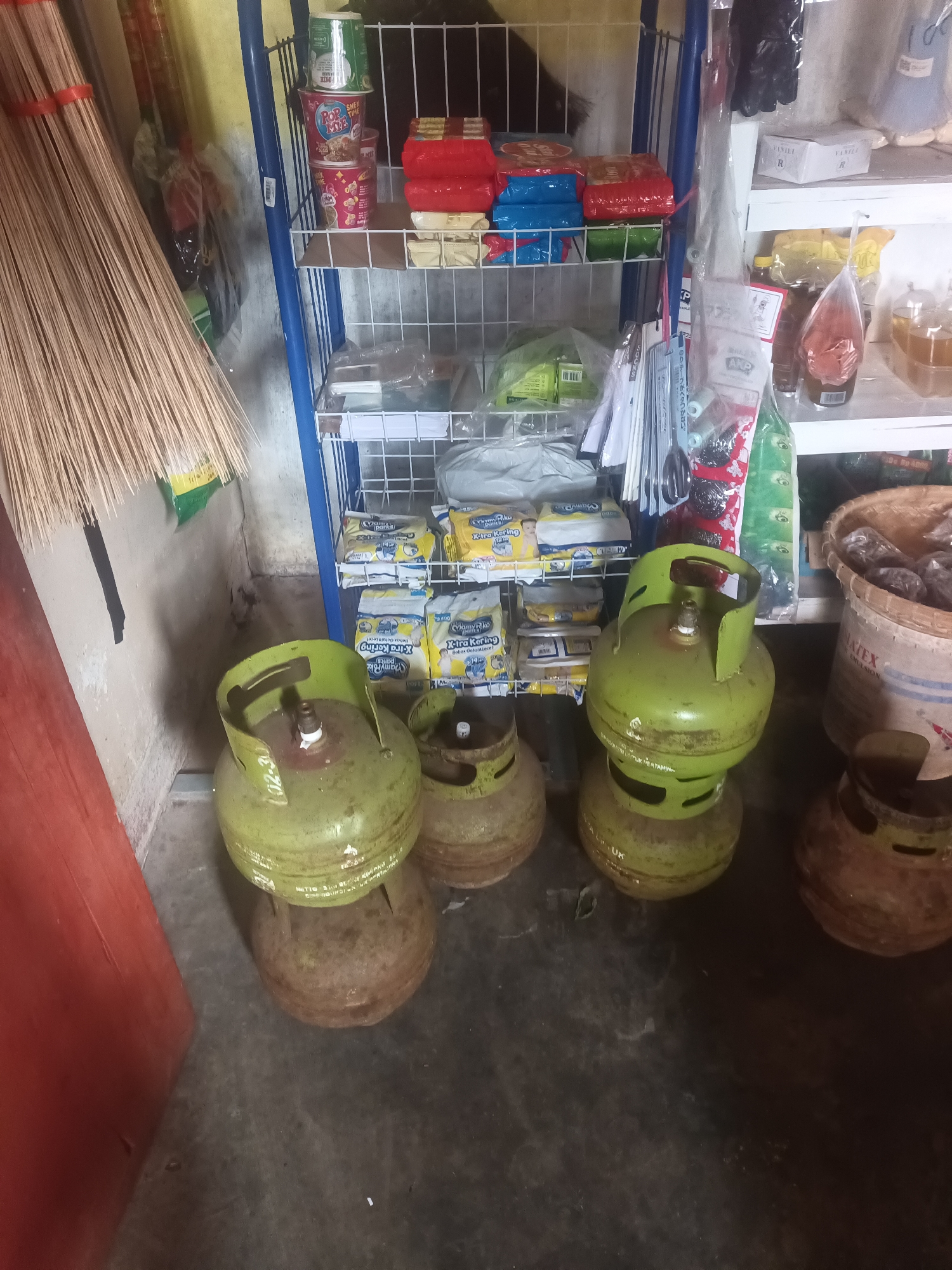 The image depicts an indoor scene, likely a small shop or storage area, filled with various items. In the foreground, three green propane tanks are prominently displayed, two of which are stacked. The tanks appear to be used or older, with some visible rust and dirt. To the left, a large bundle of brooms or similar cleaning tools leans against a wall. Behind the brooms, a section of a blue metal shelving unit is visible, holding several packaged goods, including what appear to be snacks and diapers. The shelves also contain some plastic-wrapped items and a large bucket. Further back, more shelves are visible, holding various packaged food items and containers of liquids. The environment appears somewhat cluttered and utilitarian, suggesting a functional space rather than a retail display. The lighting is ambient, coming from an unseen source, casting some shadows. The exact location in Terbanggi Marga, Indonesia cannot be determined from the image alone, but the presence of local Indonesian products on the shelves, like snacks and diapers, might suggest a local shop.