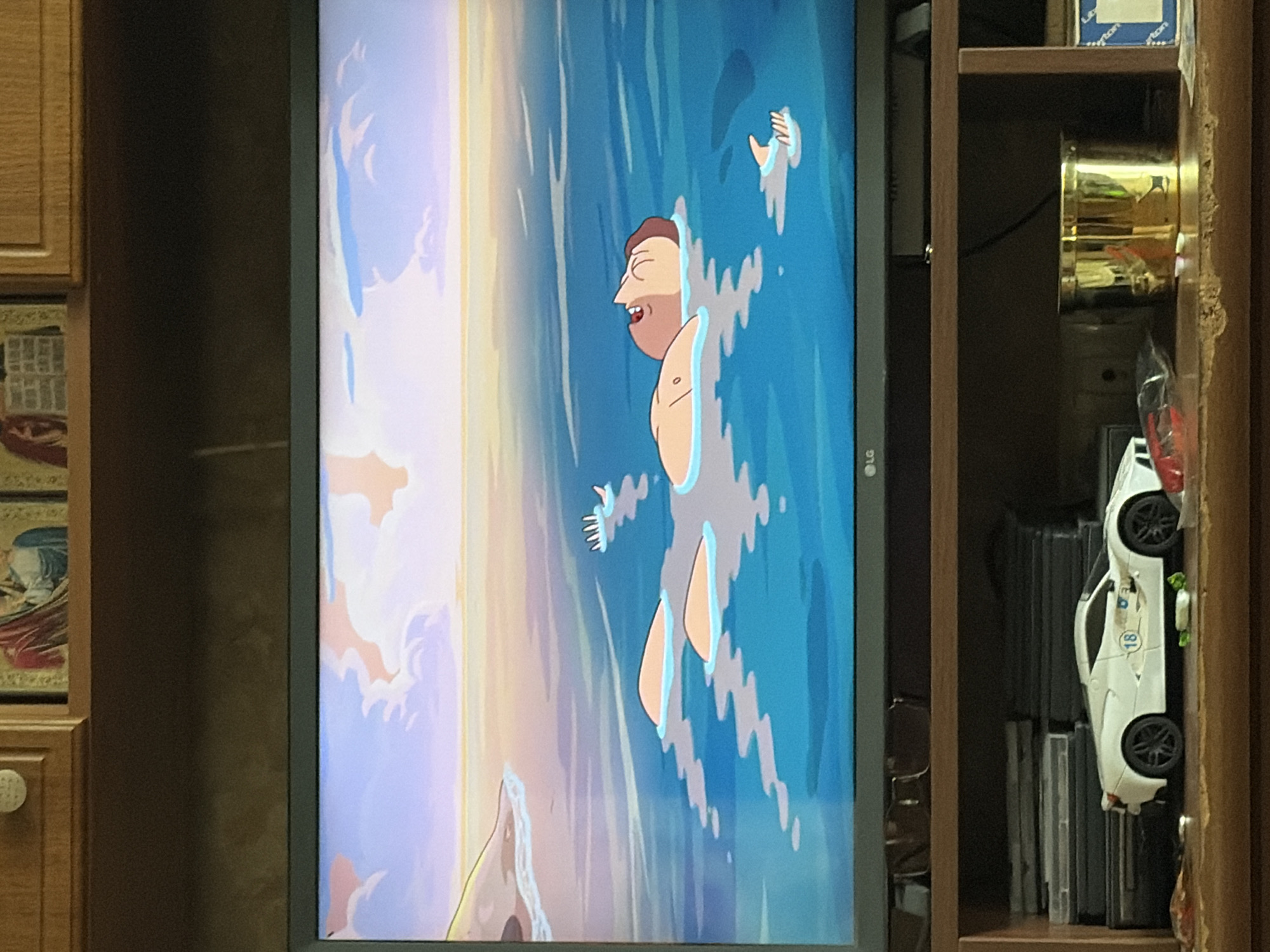 The image displays a television screen showing an animated scene of a person floating on their back in the ocean. The person's face is tilted upwards, with their mouth slightly open. Several light orange objects, possibly buoys or floating debris, are scattered around the person in the water. In the background, a gentle coastline is visible with soft, pastel-colored clouds in the sky. Below the television, a wooden shelf holds a white toy car with the number 18 on its side, a stack of DVDs or games, and a gold cylindrical container. The scene appears to be a still from an animated show or movie.