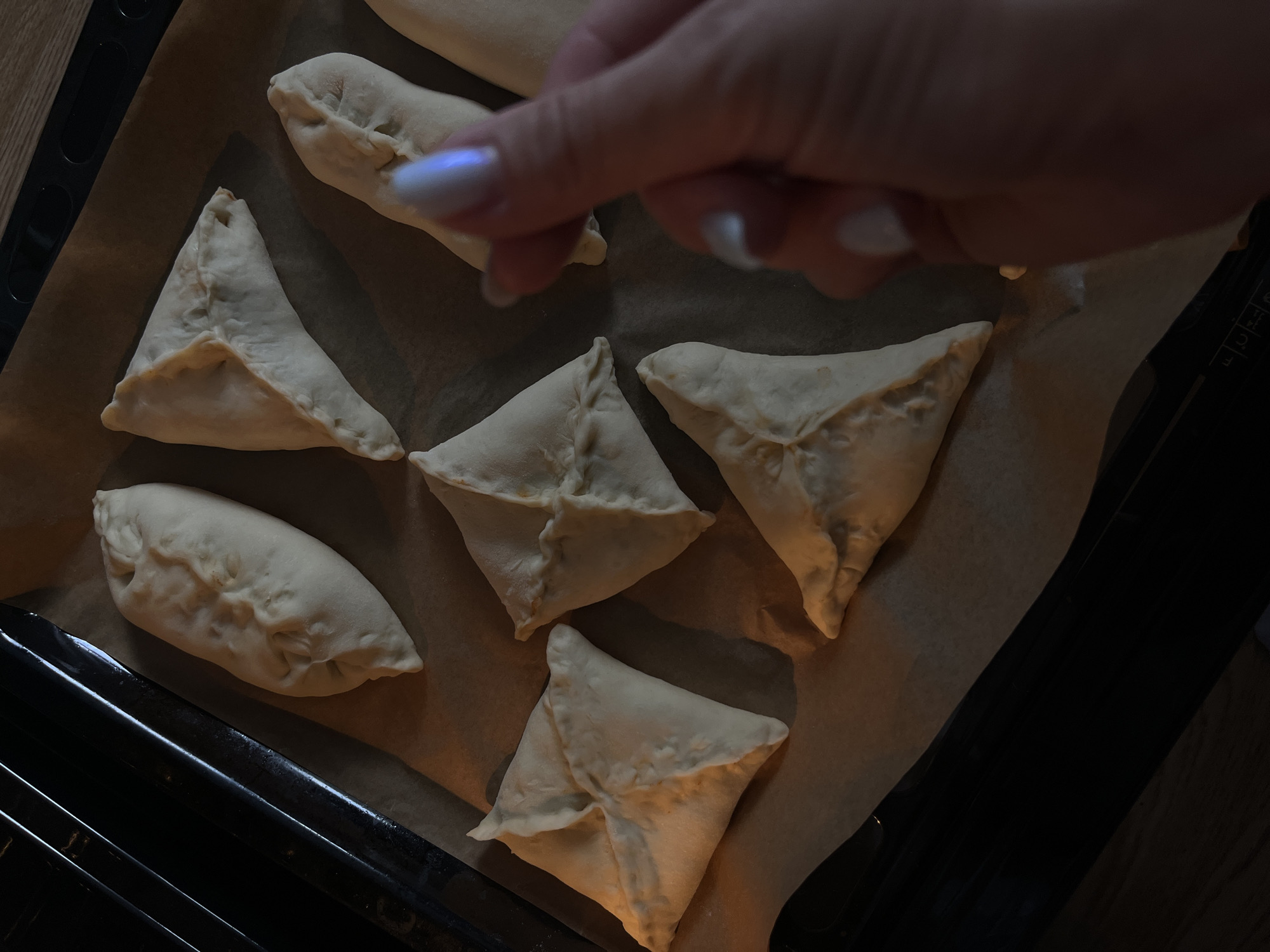 The image depicts a baking sheet lined with parchment paper, holding six pastries. Four pastries are triangular, with pointed edges, while two are oblong and crescent-shaped. A hand with manicured nails is reaching towards the pastries. The baking sheet is situated inside an oven. The location is undetermined as there is no way to tell this is Shushary, Russia.