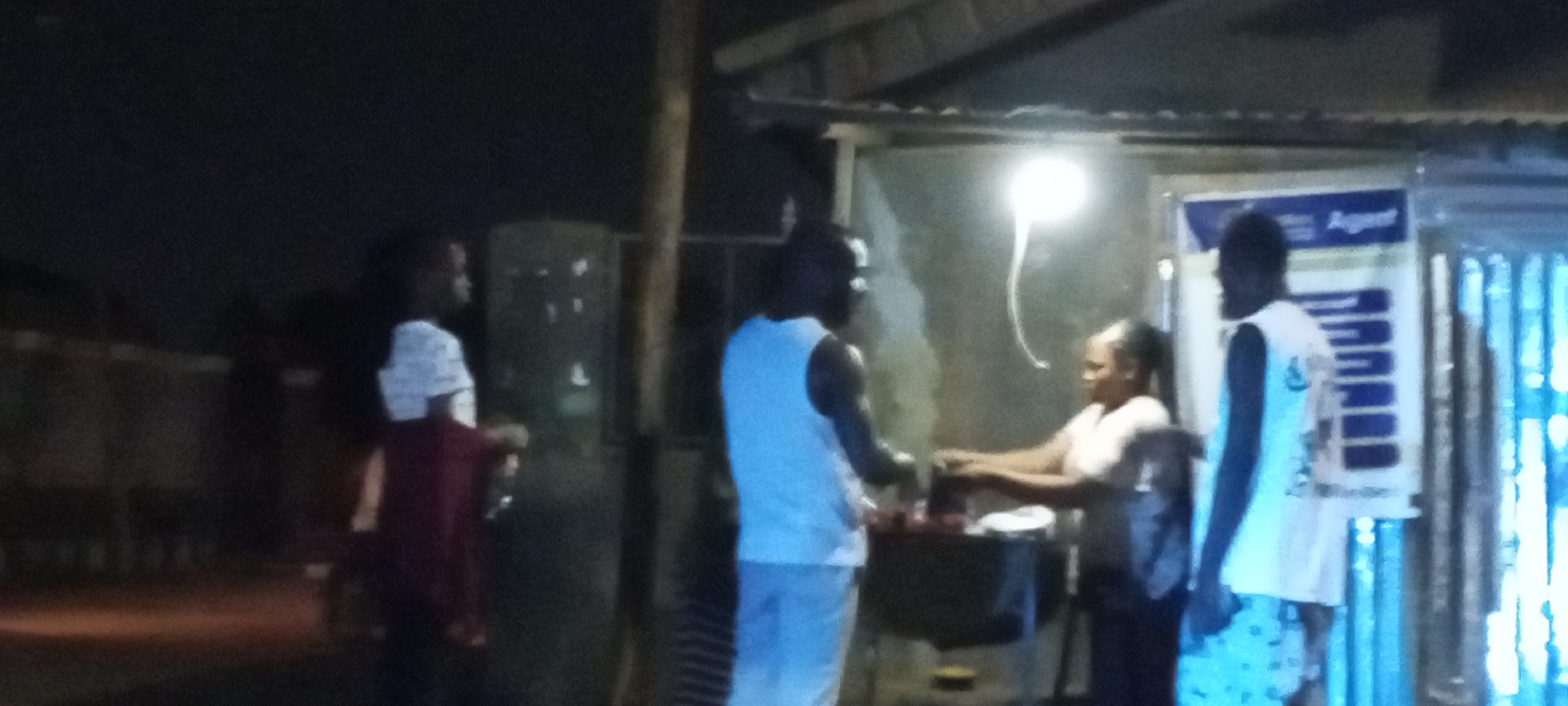 The image depicts an outdoor scene at night in Sabon Tasha, Nigeria. A woman is standing behind a dark-colored table or cart, apparently serving food to a man in a light-colored tank top and pants. Another man stands to the right, also in light clothing, near a vertical sign that reads "Agent" at the top. To the left, another individual in a light shirt and red jacket stands further back. An overhead light illuminates the area. The setting appears to be a roadside food stall under a corrugated metal awning or roof. The background is dark and indistinct.