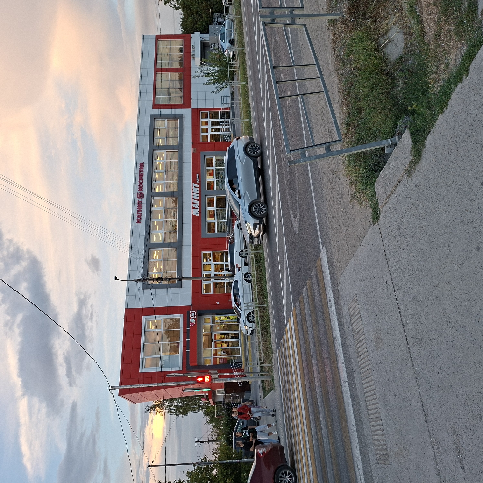 The image captures a street scene in Anapskaya, Russia, in the late afternoon or early evening, indicated by the soft, warm lighting and cloud formations. The focal point is a modern, two-story retail building with a distinctive red and grey facade. Prominently displayed on the building are the signs "МАГНИТ КОСМЕТИК" and "МАГНИТ у дома," identifying it as a Magnit Cosmetics and a Magnit convenience store. The building has large, multi-paned windows on both levels, reflecting the sky and surrounding trees.
In the foreground, a pedestrian crossing with yellow and white stripes is visible on a paved road. A silver SUV is driving across the intersection. To the left of the crossing, a traffic light displays a red upward arrow and the number "86." A group of four individuals is standing on the sidewalk near the entrance of the store, with one person wearing a red jacket and another in a black t-shirt. A dark-colored car is parked behind them. A red sedan is partially visible in the bottom left corner of the image.
The environment includes trees and vegetation along the roadside and behind the building, and a metal safety barrier is present on the right side of the image. The sky is partly cloudy, with hues of pink and orange suggesting the setting sun. Electrical wires are visible crisscrossing the sky above the building. The overall impression is one of everyday urban life in a Russian town.
